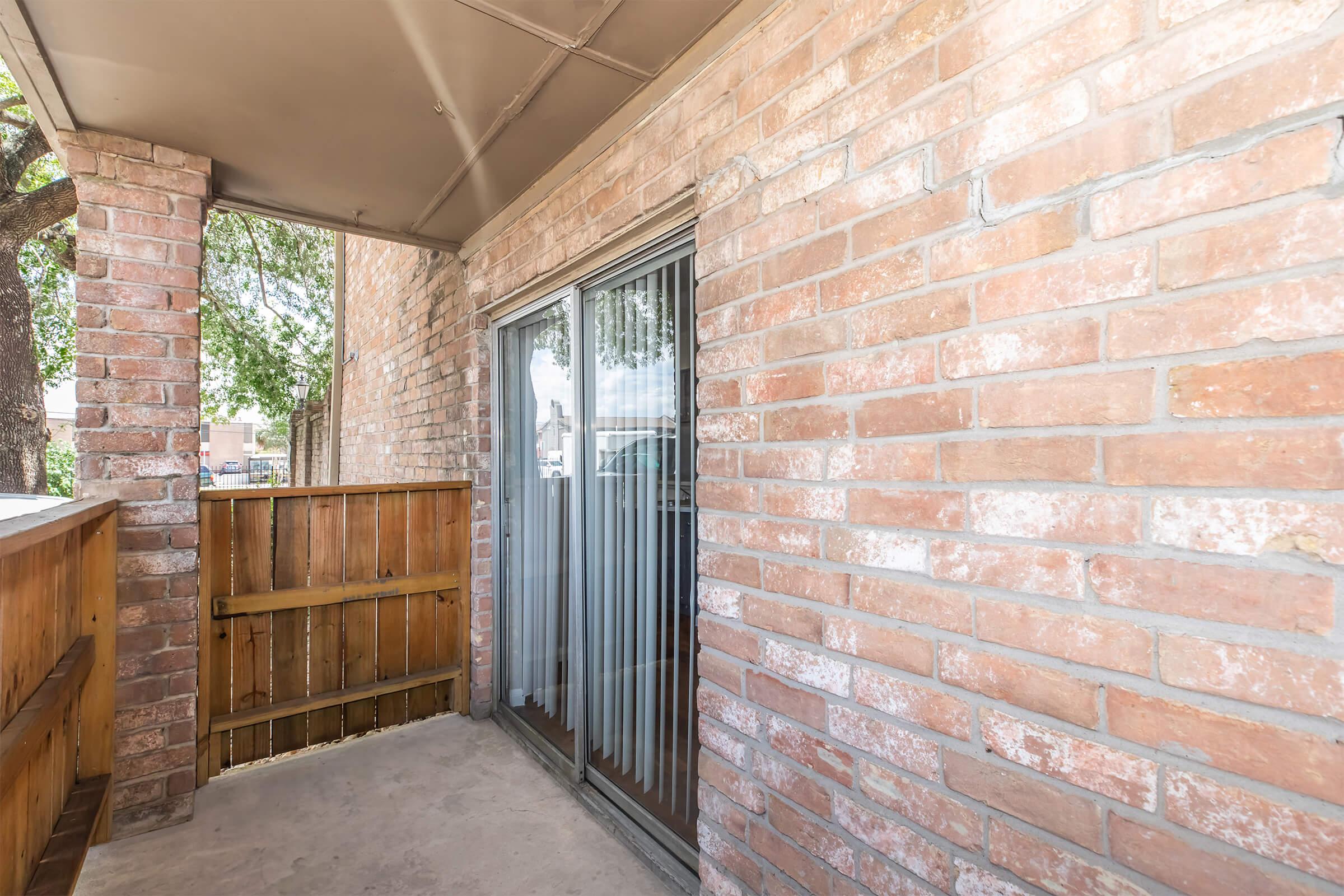 A small balcony area featuring brick walls, a sliding glass door with vertical blinds, and a wooden fence. The space is well-lit with natural light coming through the door. There are trees visible outside, adding to the outdoor ambiance.