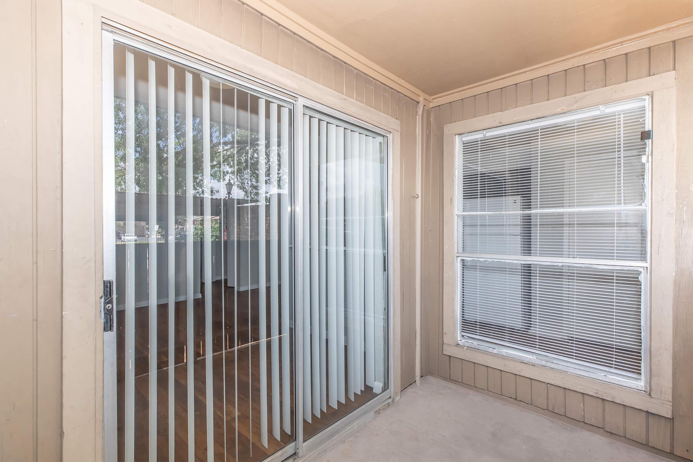 A view of a covered patio or balcony featuring glass sliding doors with vertical blinds. There is a window with horizontal blinds on the right side. The walls are painted in a light brown color, and the flooring is wooden. Natural light enters from the outside, illuminating the space.