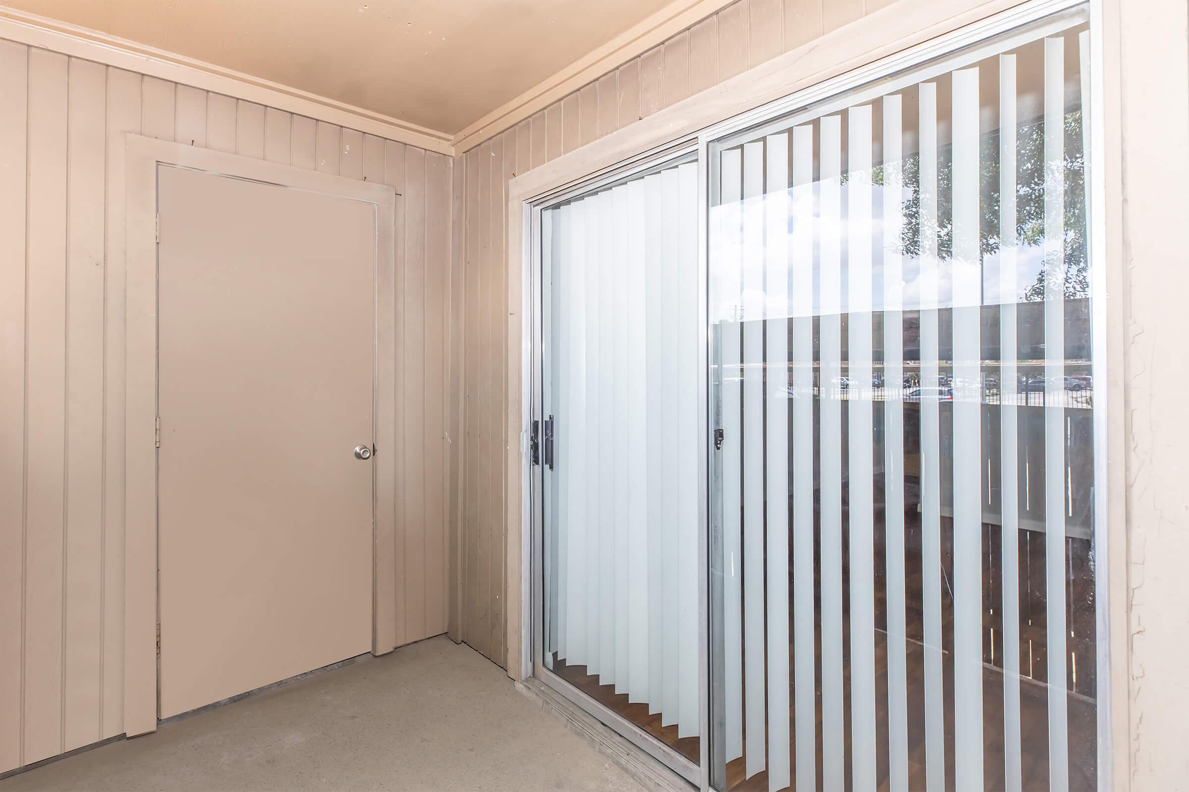 An interior view of a room featuring a wall with vertical blinds covering a sliding glass door. There is a plain door to the left, and the walls are painted light beige. Sunlight filters through the blinds, creating a bright and airy atmosphere. The floor is plain concrete.