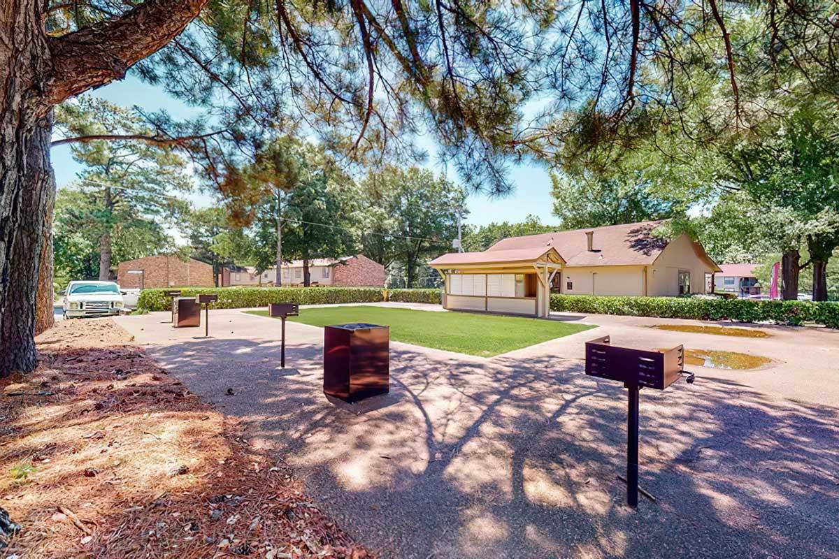 A landscaped outdoor area featuring a small building and several black informational displays on pedestals. Surrounding the space are trees, providing shade and a natural atmosphere. A grassy lawn is visible in the center, and a parking area can be seen in the background.