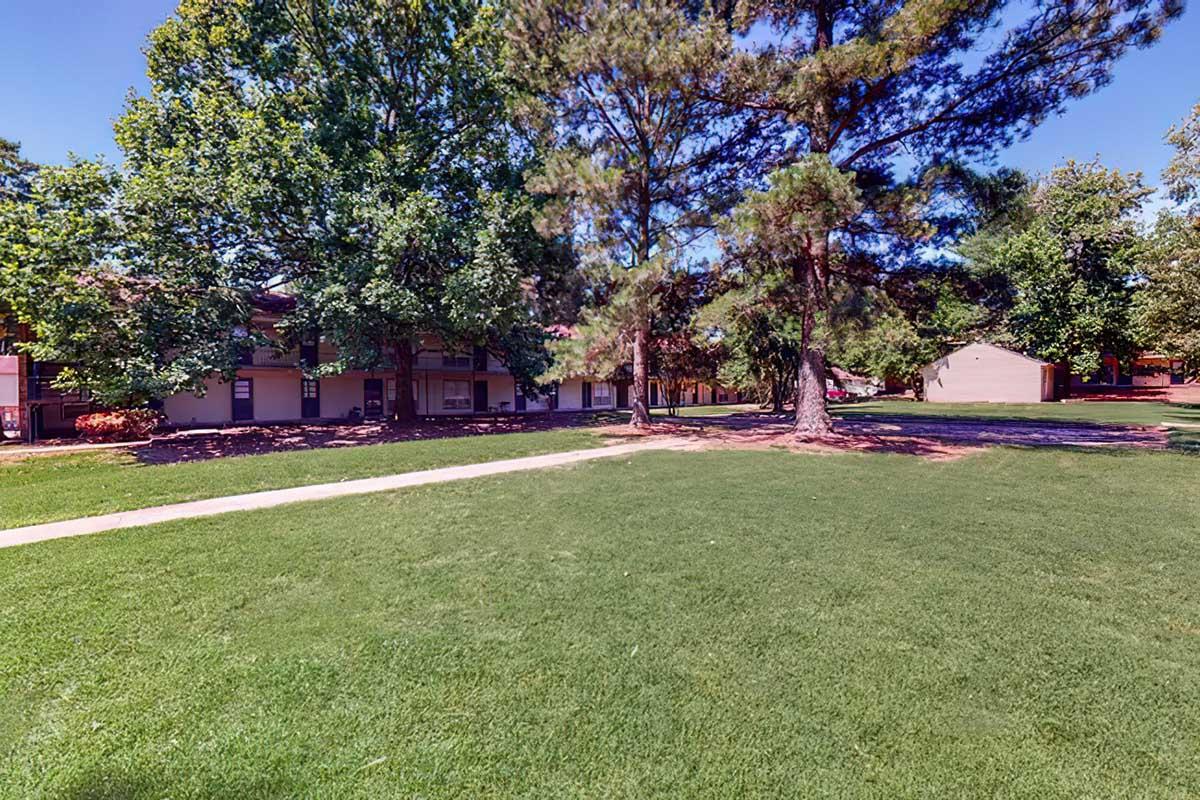 A grassy area with trees in the foreground and buildings in the background under a clear blue sky. The scene is bright and depicts a peaceful outdoor setting, suitable for relaxation or recreational activities.