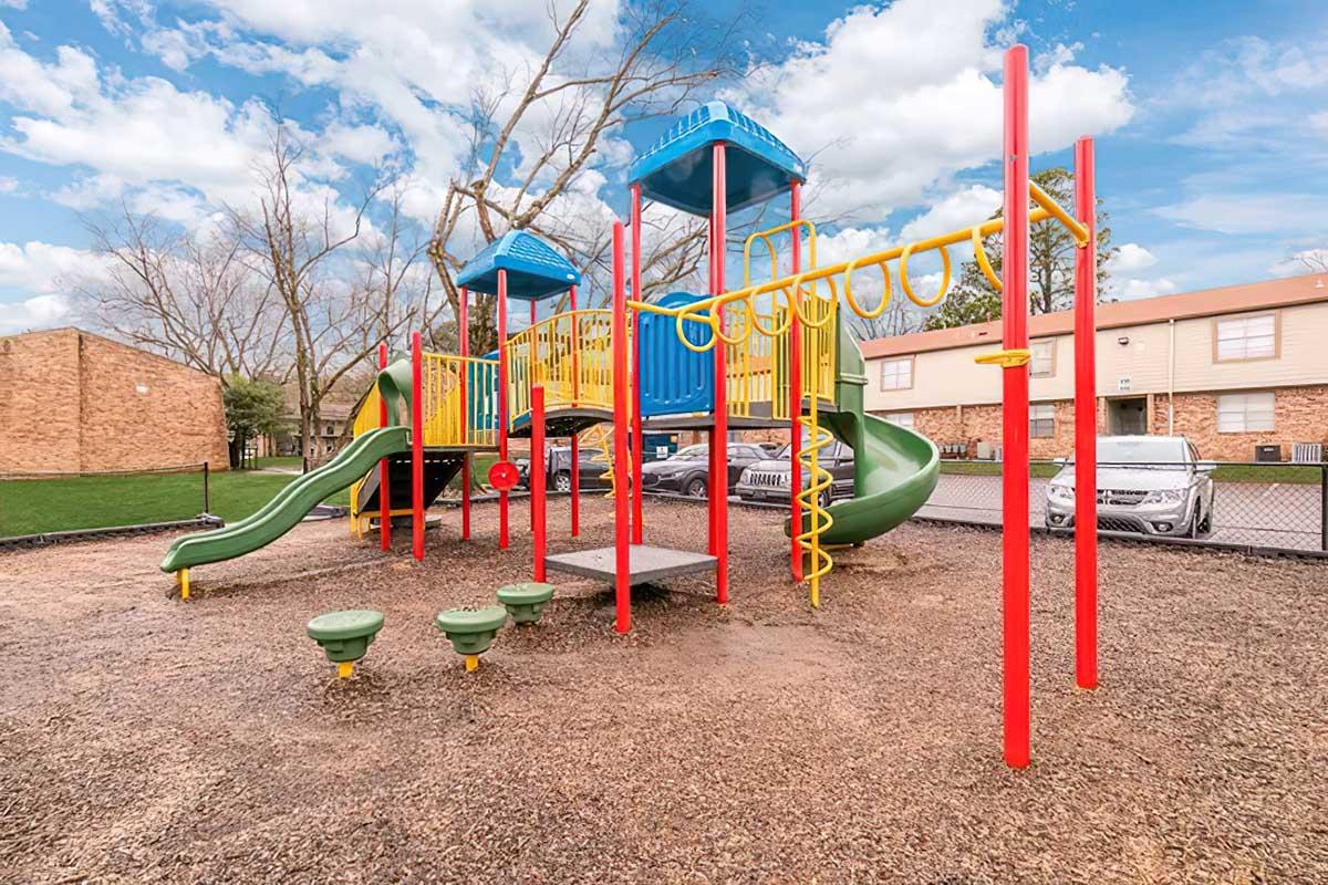 A colorful playground featuring slides, climbing structures, and monkey bars, surrounded by a fenced area with grassy patches and residential buildings in the background. The sky is blue with some clouds.