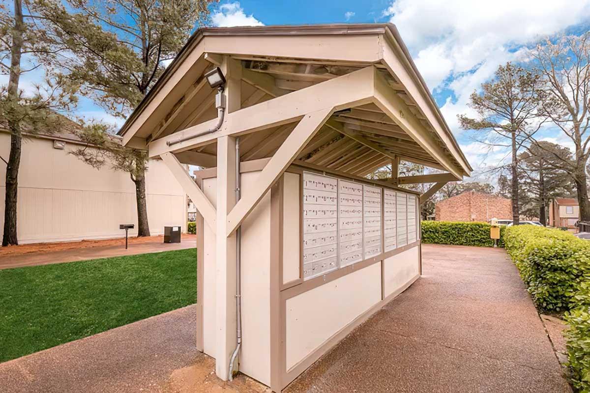 A postal kiosk with multiple mailbox compartments, situated under a peaked roof. The structure is located in a grassy area surrounded by trees, with a pathway leading up to it. The sky is partly cloudy, adding to a pleasant outdoor setting.