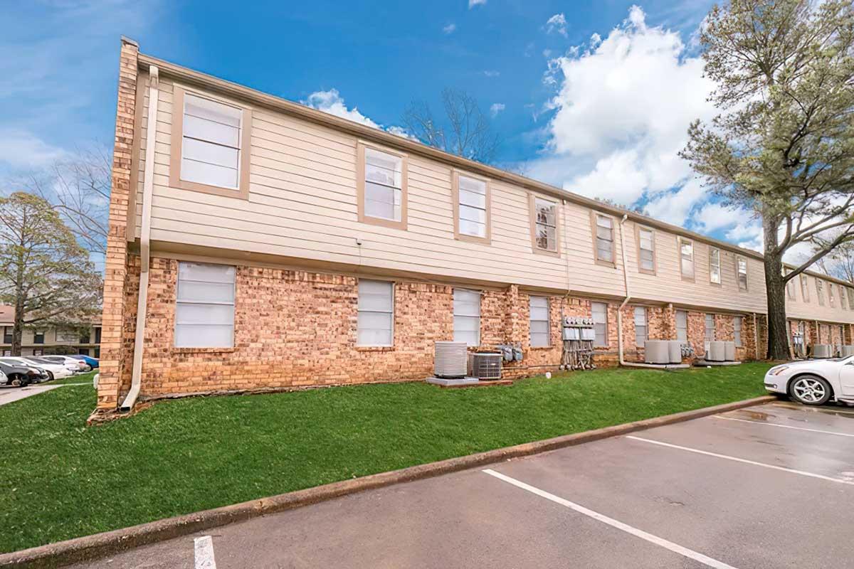 A residential building featuring a brick and siding exterior, with multiple windows and air conditioning units. The structure is set on a grassy area, adjacent to a parking lot with a white car parked nearby. The scene is set under a partly cloudy blue sky.