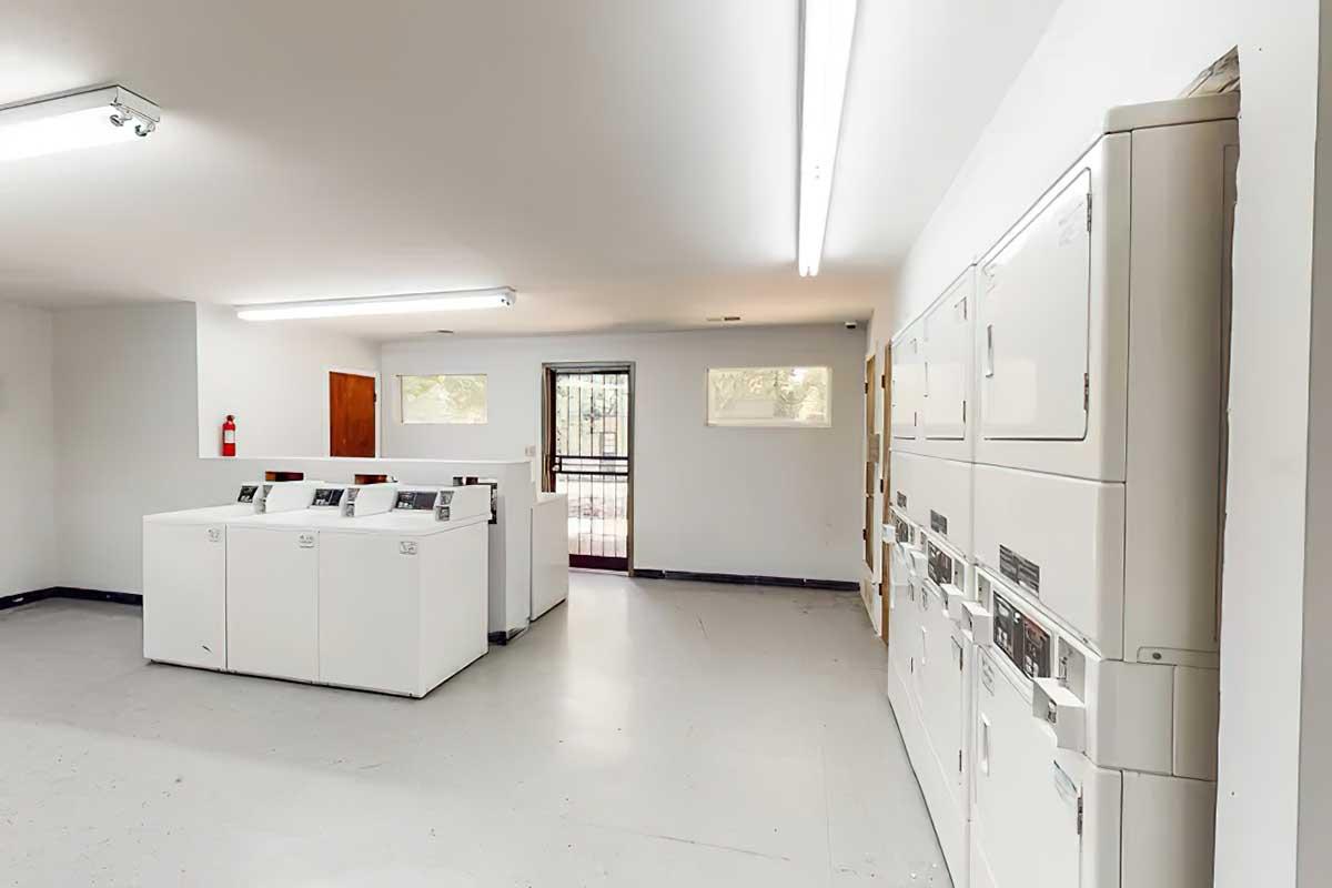 A clean, well-lit laundry room featuring several white washing machines and dryers arranged in a row. There are windows allowing natural light, a fire extinguisher mounted on the wall, and a door leading outside. The floor is light-colored and the overall atmosphere is tidy and organized.