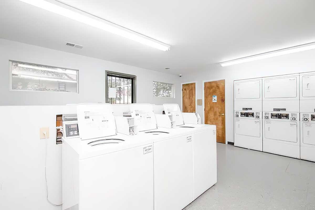 A bright, clean laundry room featuring several white washing machines and dryers lined against the wall. The space has white walls and ceilings, fluorescent lighting, and a window allowing natural light. There are multiple machines available for use, indicating a shared laundry facility.
