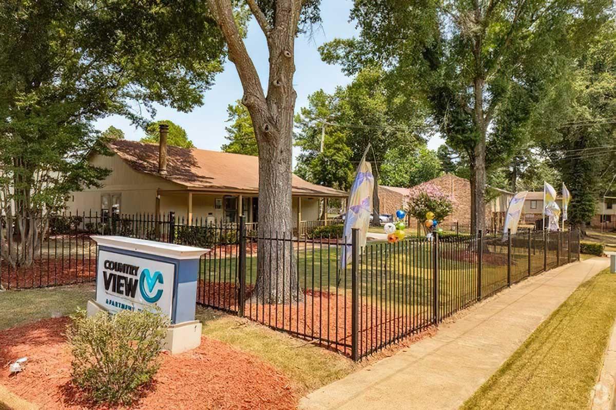 A well-maintained property with a sign reading "Country View," surrounded by a black wrought iron fence. The yard features manicured grass, colorful decorations, and a few flags, set against a backdrop of tall trees and blue skies. The house has a warm, inviting exterior with a brown roof.