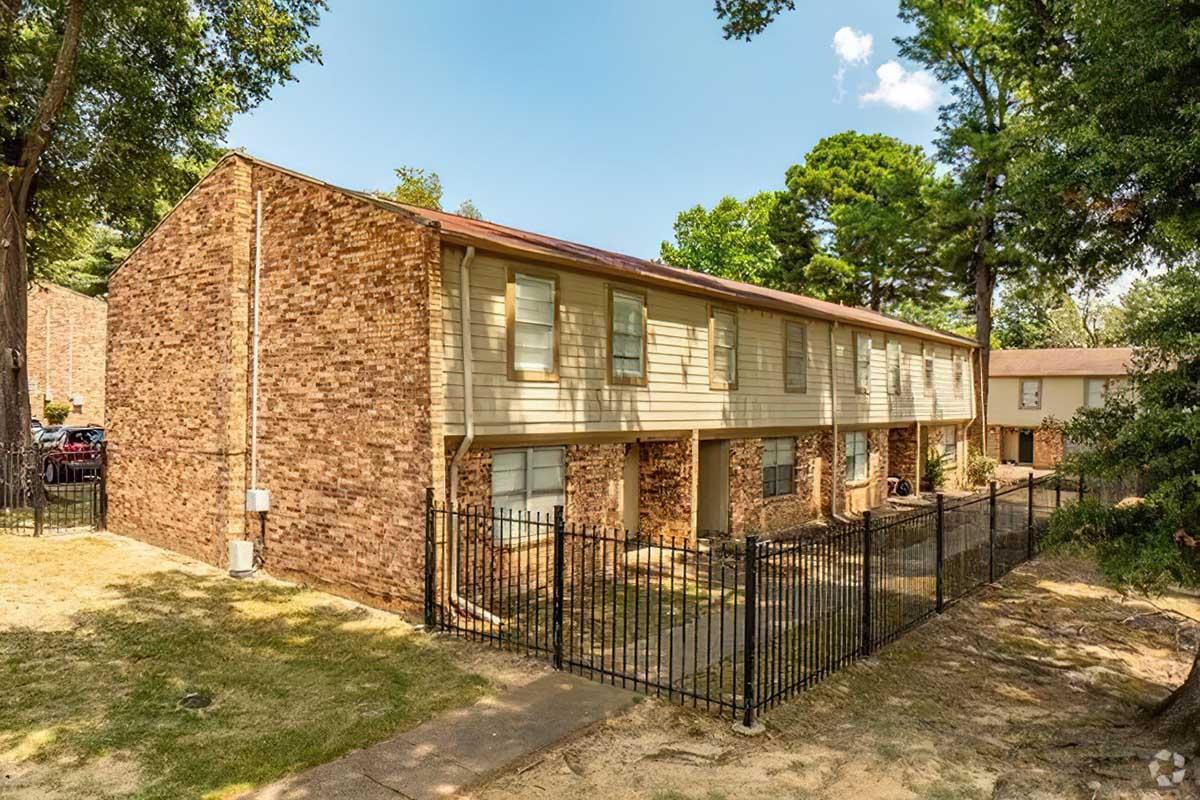 A two-story brick apartment building with a fenced yard, surrounded by trees and greenery. The building features multiple windows and a sloped roof, with a pathway leading to the entrance. Bright blue skies are visible in the background.