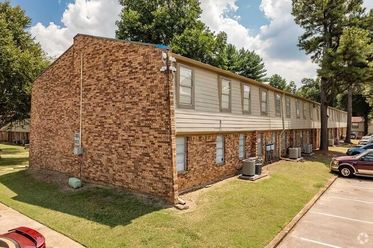 A brick apartment building with multiple units, featuring air conditioning units on the exterior walls. The building is surrounded by green grass and trees, with parking spaces nearby. The sky is partly cloudy, creating a bright atmosphere.