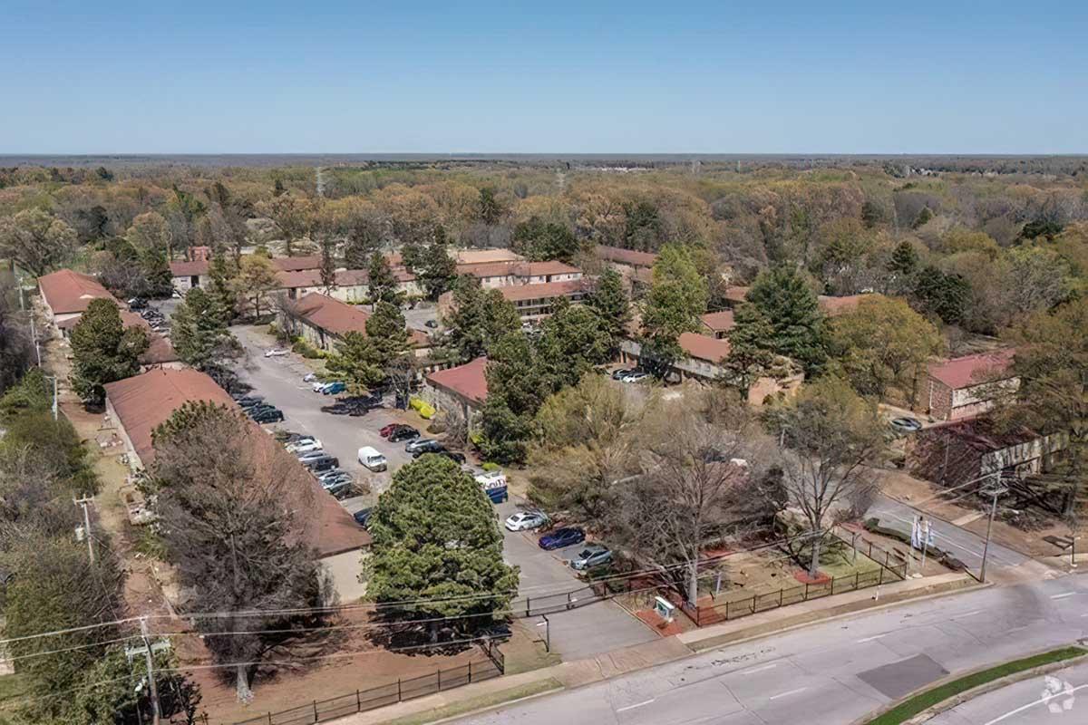 Aerial view of a residential area featuring low-rise buildings surrounded by trees. The parking lot shows various vehicles, and the landscape includes green spaces and pathways, with power lines visible along the road at the bottom. The sky is clear and blue, indicating a sunny day.