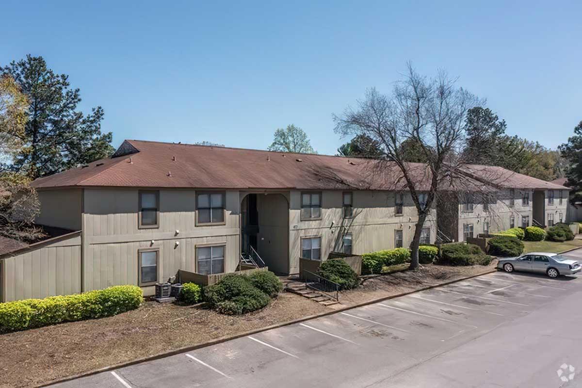 A row of two-story apartment buildings with a light brown exterior, surrounded by manicured shrubs. The parking lot in front has a few parked cars and is lined with trees. The sky is clear and blue, indicating a sunny day.
