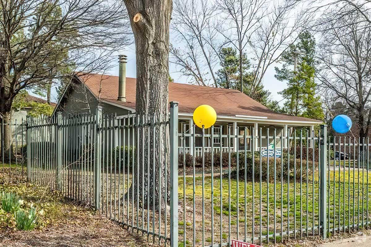 A single-story house with a brown roof and large windows is surrounded by a green fence. Two balloons, one yellow and one blue, are attached to the fence. Trees are visible in the background, and the yard appears well-maintained with grass and some plants.