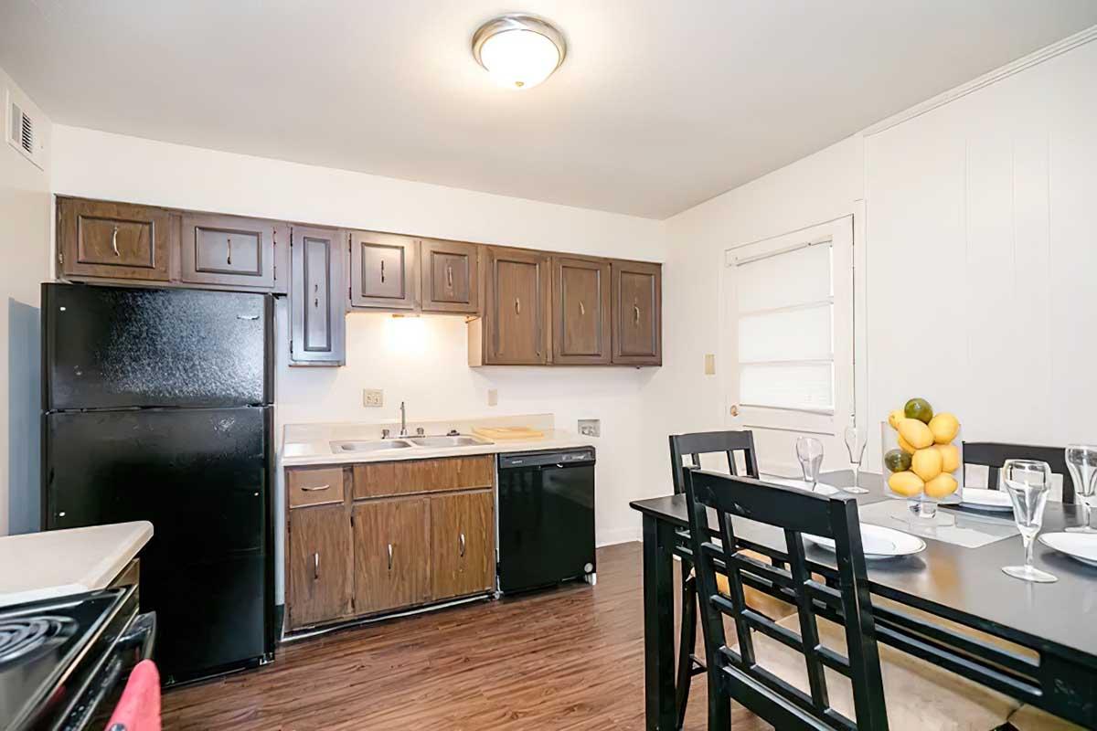 A small kitchen featuring dark brown wooden cabinets, a black refrigerator, and a black dishwasher. There's a sink with a countertop space and a table set for two with plates, glasses, and a bowl of fruit, including lemons and oranges. The floor has a wood finish, and the walls are light-colored.