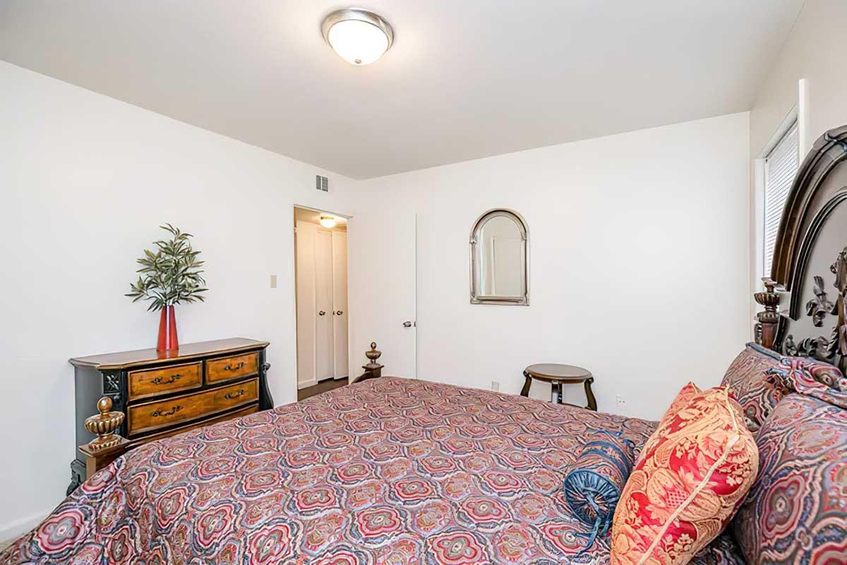 A cozy bedroom featuring a decorative bed with a patterned cover, bright pillows, a nightstand, an ornate dresser, and a small table. A potted plant adds a touch of greenery. The walls are painted white, and there’s a mirror on the wall, contributing to a welcoming atmosphere.