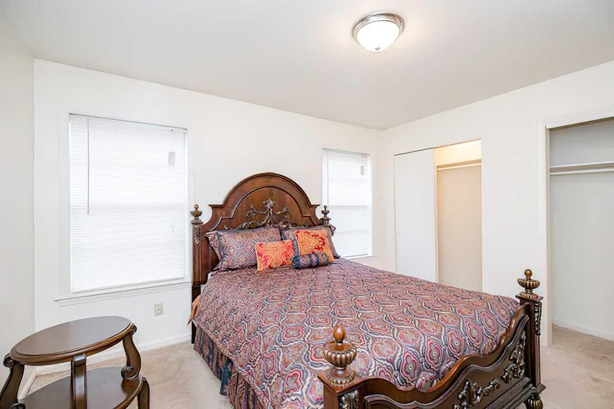 A cozy bedroom featuring a decorative wooden bed with a patterned blanket and vibrant pillows. There are two windows with blinds, a small round bedside table, and a spacious closet. The walls are painted white, creating a bright and inviting atmosphere.