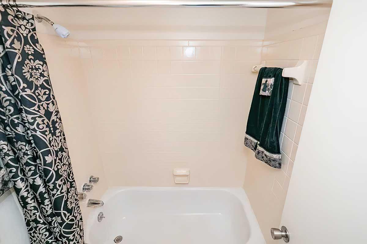 A clean, white bathtub with silver faucets and a showerhead. A patterned black and white shower curtain hangs on the side, and a green towel is neatly placed on a towel rack nearby. The bathroom has light-colored tiled walls and a simple aesthetic.