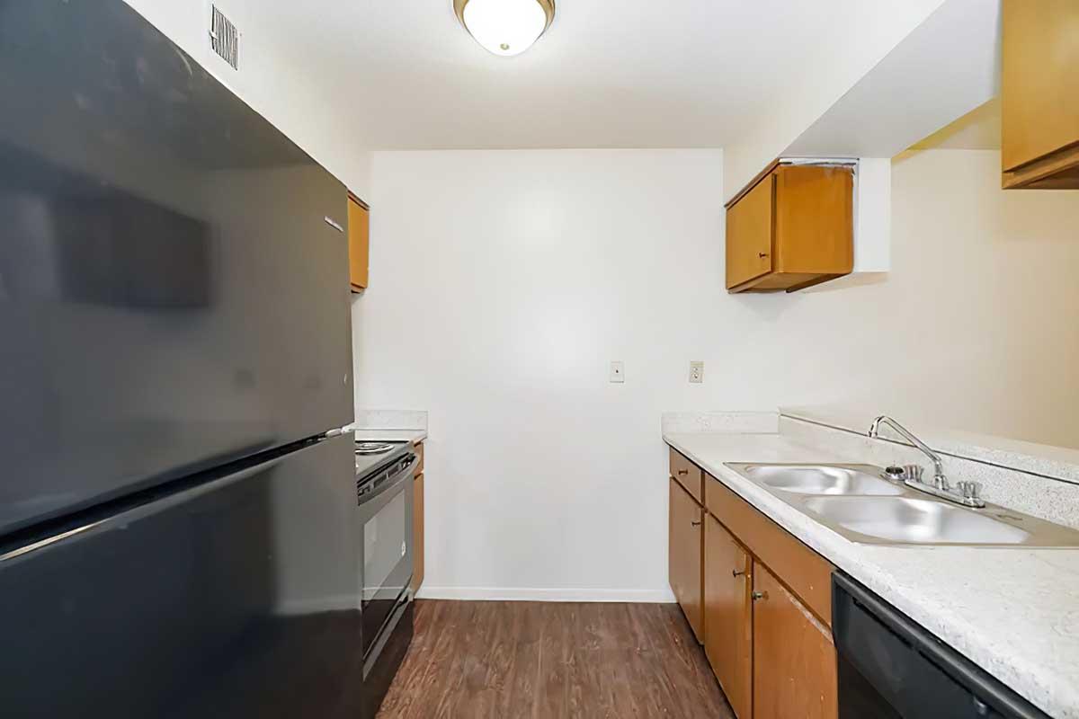 A kitchen interior featuring a black refrigerator on the left, a black stove, and two stainless steel sinks on a white countertop. Wooden cabinets are mounted above the counters, and the walls are painted white. The flooring is dark wood laminate, creating a modern and clean look.
