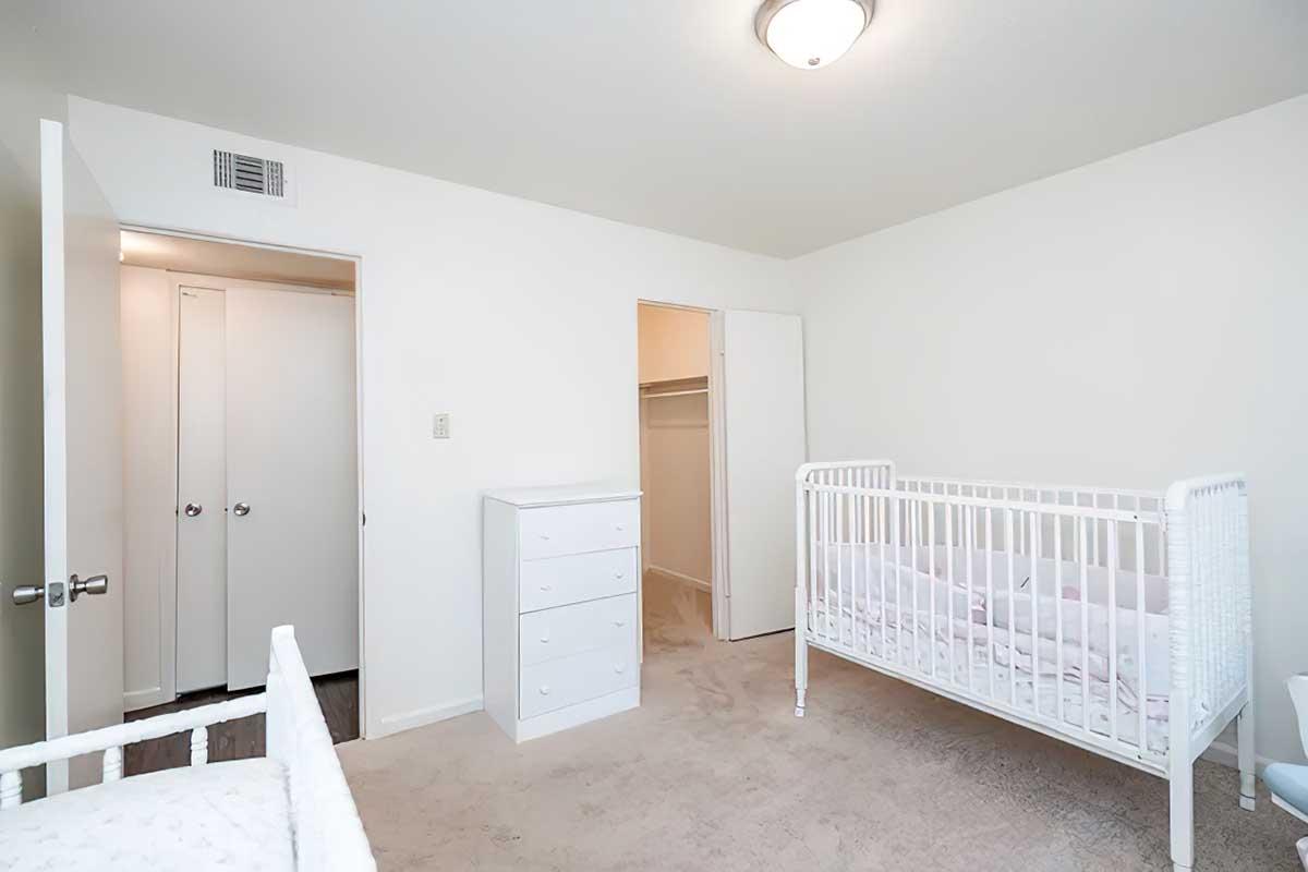 A bright, empty nursery featuring a white crib and a white dresser against light-colored walls. There is a closet door partially open, revealing a simple design. The carpeted floor adds warmth to the space, which is designed for a child. The room is uncluttered and inviting.
