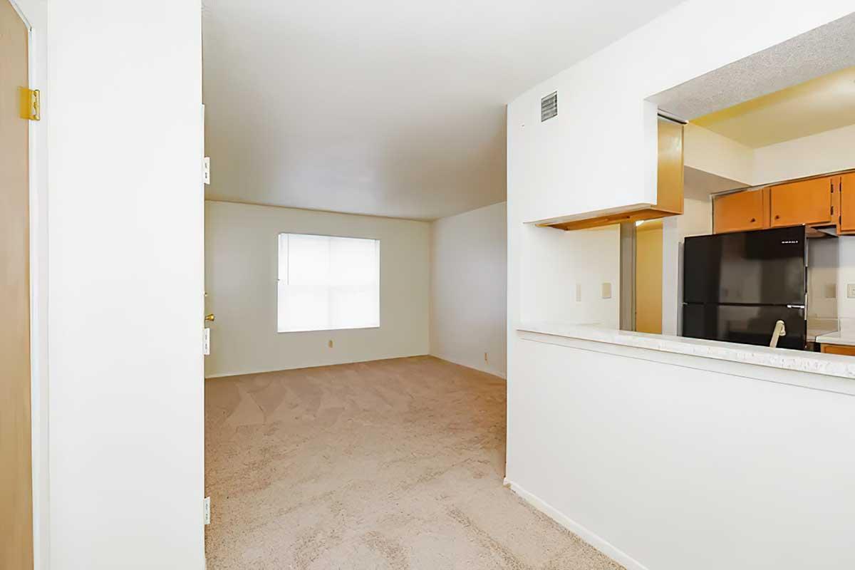 A bright, carpeted living area with a window letting in natural light, adjoining a kitchen with wooden cabinets. An open layout features a small bar area separating the kitchen from the living space. The walls are painted white, creating a clean and airy atmosphere.