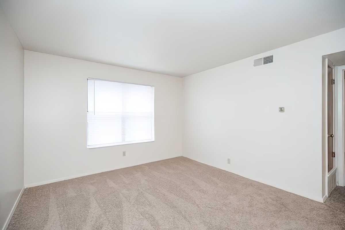 A spacious, empty room with light-colored walls and beige carpet. A large window with closed blinds lets in natural light, and there is a door on the right side leading to another space. The overall atmosphere is neutral and minimalistic, providing a blank canvas for potential decoration.