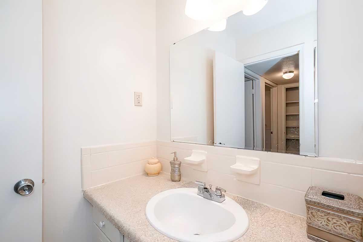A clean, modern bathroom featuring a white sink with a chrome faucet, a large mirror above it, and beige countertops. There are two soap dispensers mounted on the wall, a decorative jar on the counter, and a silver container next to the sink. Light-colored walls and a door leading to another room are visible in the background.