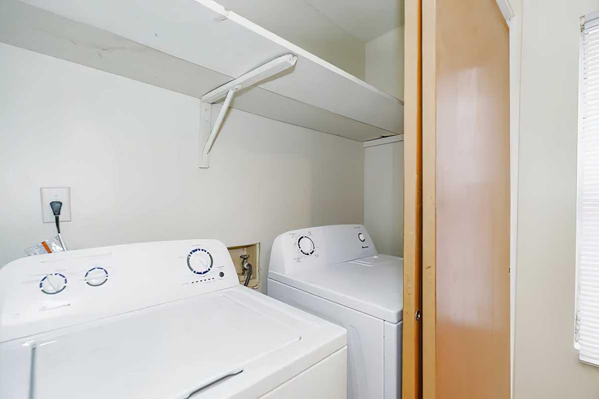 A small laundry area featuring two white washing machines placed side by side, with a shelf above for storage. A door partially closed is visible on the right, and natural light filters in from a nearby window.