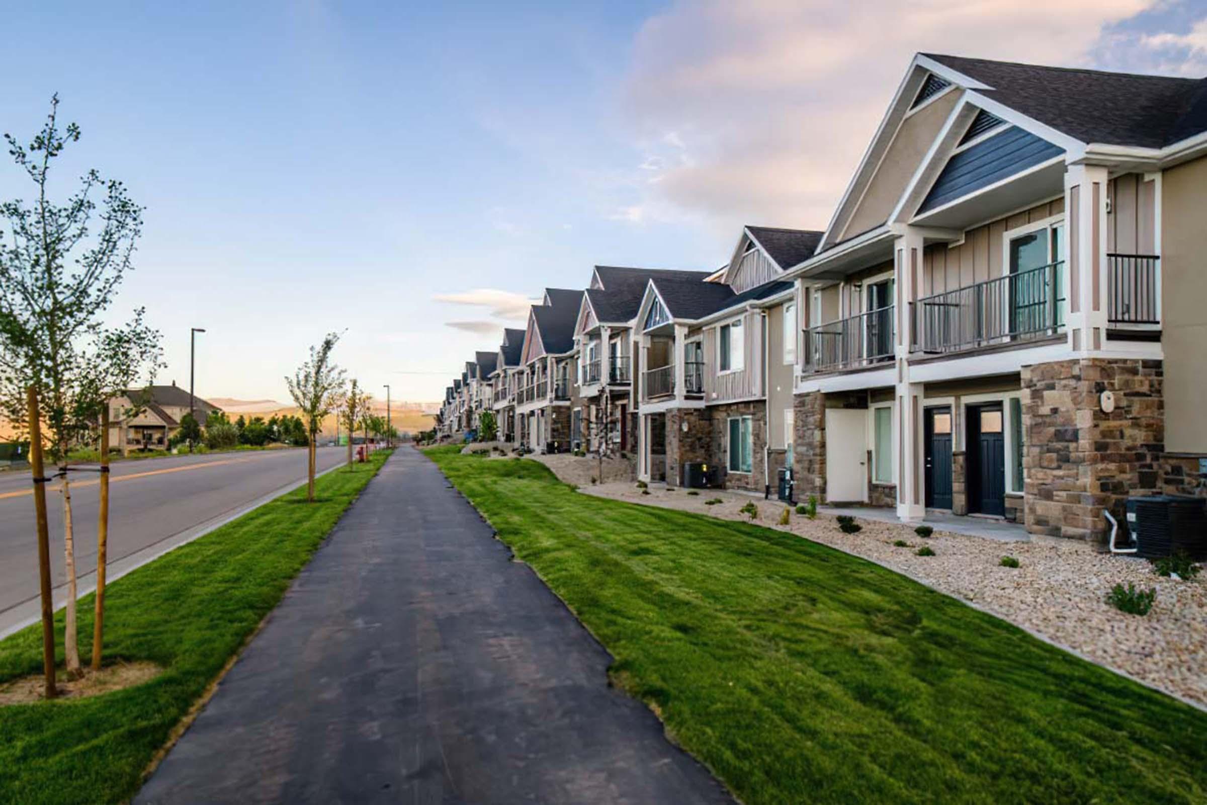 A scenic view of a modern residential street lined with well-maintained townhouses. The buildings feature a mix of stone and siding exteriors under a clear blue sky. A paved walkway runs beside a lush green lawn, enhancing the inviting atmosphere of the neighborhood.