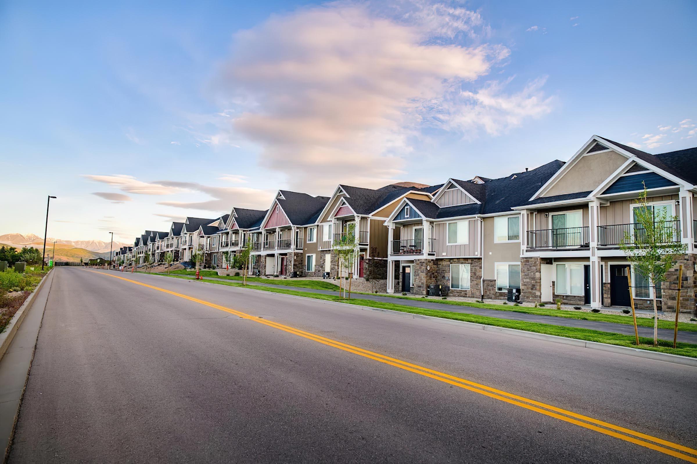 A quiet suburban street lined with modern townhouses. The houses feature a mix of architectural styles with varying colors and balconies. The scene is set against a backdrop of a clear blue sky with scattered clouds, and well-maintained grass and landscaping along the roadside.