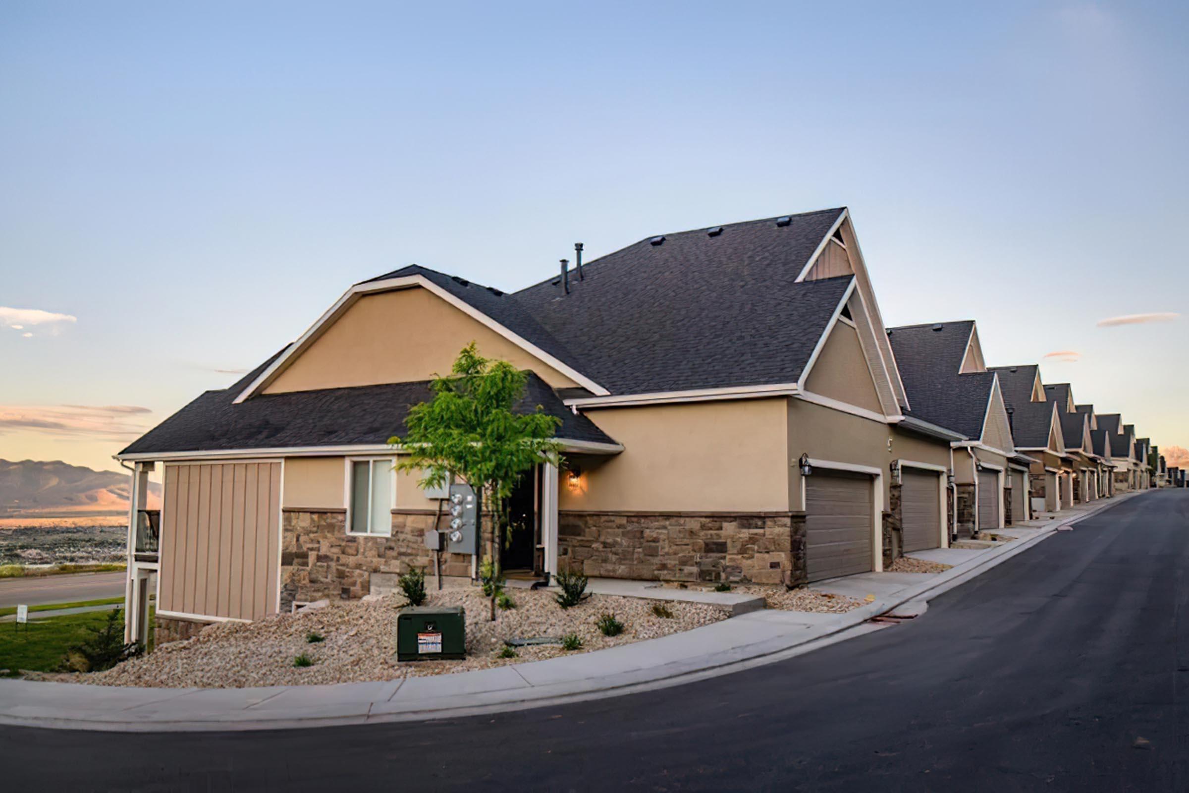 A row of modern single-family homes with stone and stucco exteriors. Each house features a sloped roof and a small front yard with landscaping. The street is paved and flanked by grass, with mountains visible in the background under a clear sky during sunset.