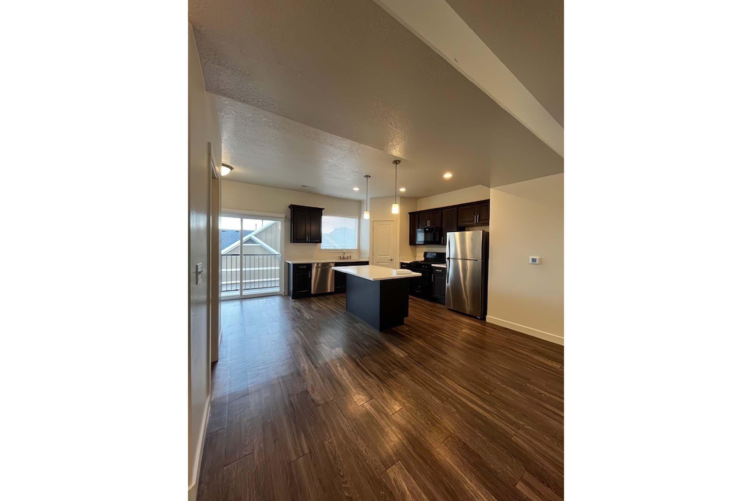 A modern kitchen with dark wooden cabinets and stainless steel appliances. There is an island in the center, and large windows allow natural light to fill the space. The flooring is a rich brown wood, and there is access to a deck visible through the sliding glass doors.