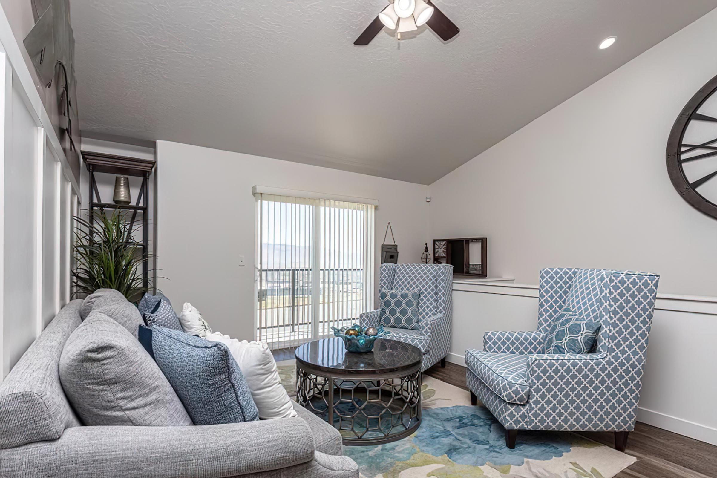 A modern living room featuring two patterned armchairs and a cozy gray sofa. A round coffee table sits in the center, adorned with decorative items. Light streams in through a sliding glass door with vertical blinds. The room is decorated with a colorful area rug and a large wall clock.