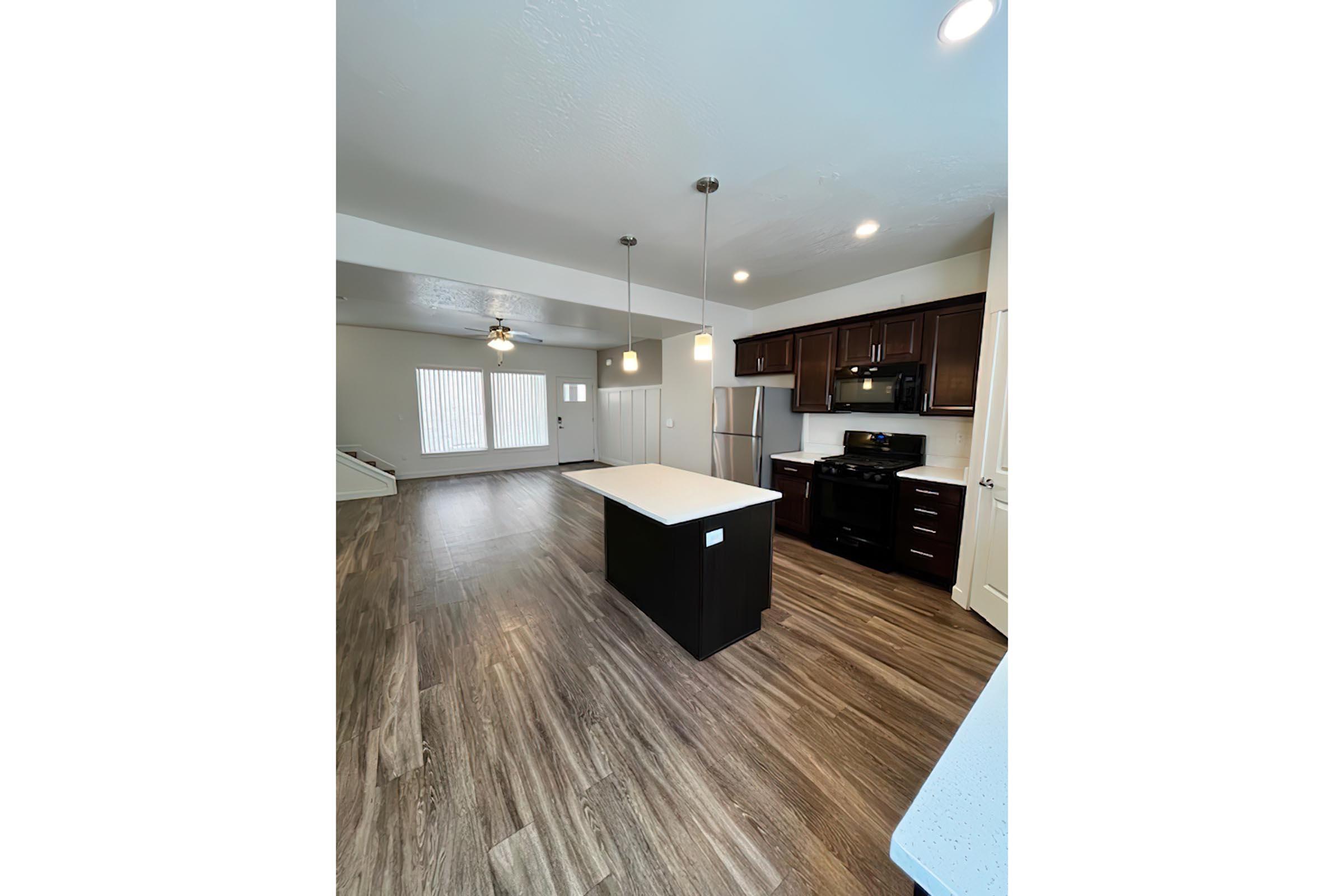 A modern kitchen with dark wooden cabinets, stainless steel appliances, and a central island with seating. The space features an open layout with a large living area, natural light from windows, and wood-style flooring. The walls are painted in a light color, enhancing the spacious feel.