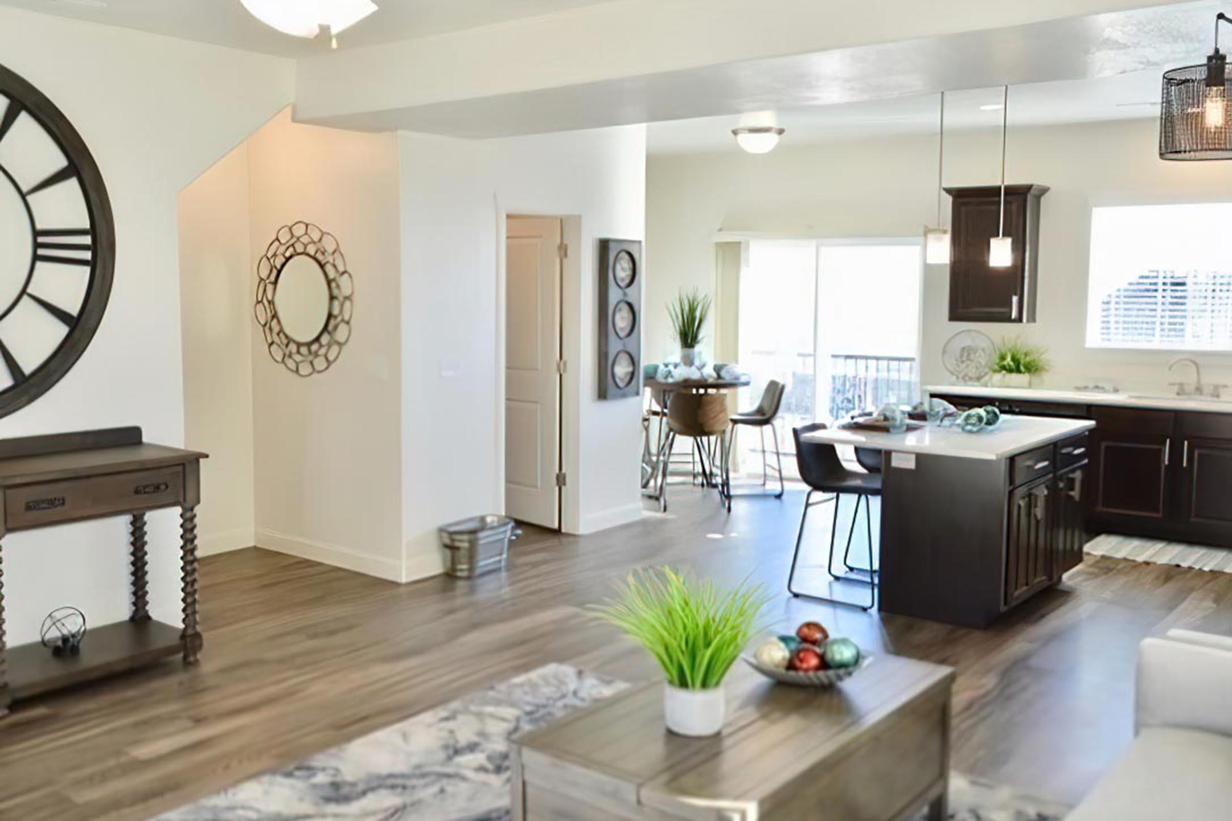 A modern, open-concept living space featuring a stylish kitchen with dark cabinets, a dining area with bar stools, and a cozy seating area. Decorative elements include a large wall clock, mirrors, and indoor plants. Natural light streams in through large windows, enhancing the contemporary ambiance.