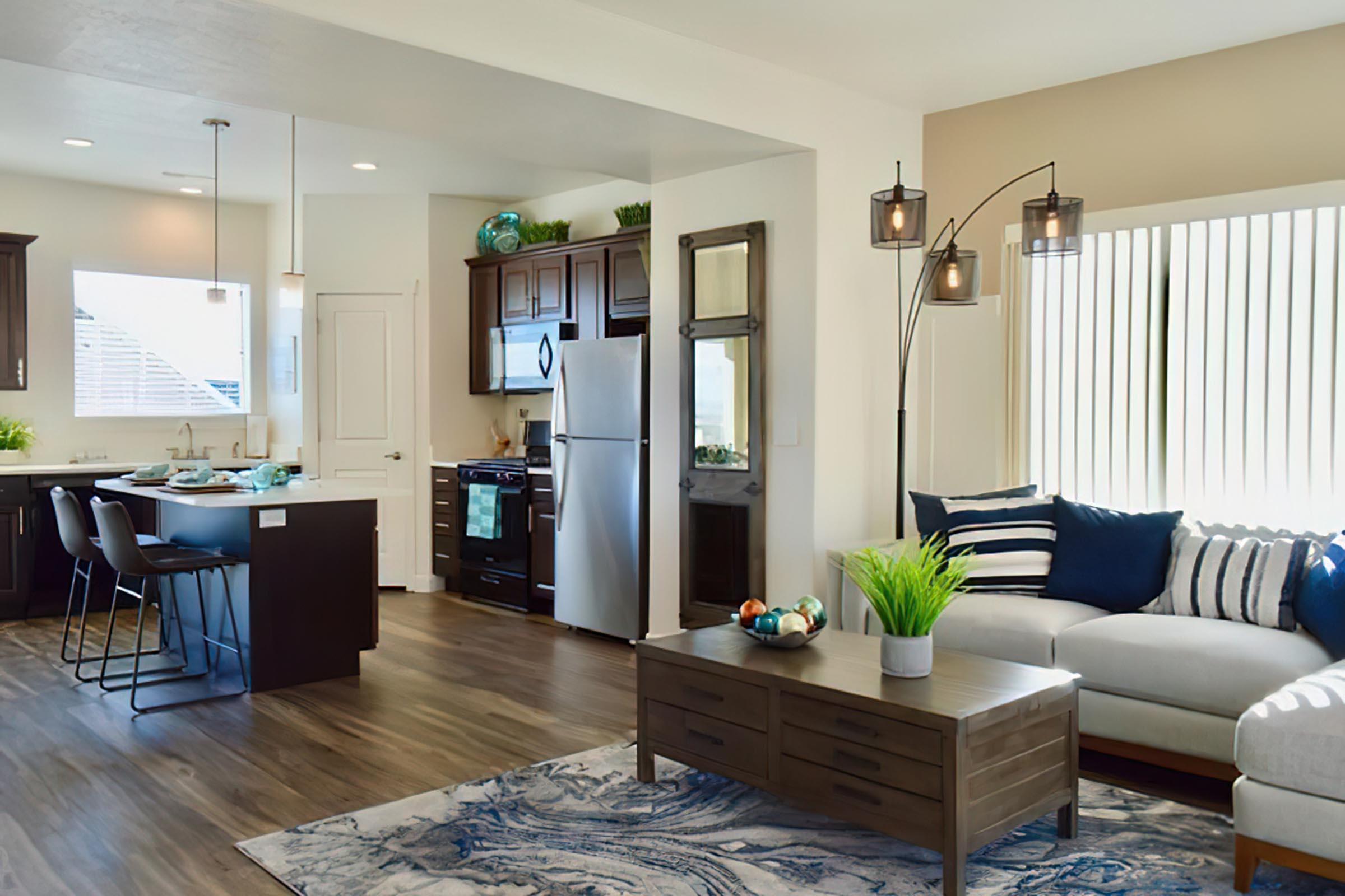 Modern open-concept living space featuring a kitchen with dark wood cabinetry, stainless steel appliances, and a light-filled living area. A comfortable sectional sofa with decorative pillows is paired with a stylish coffee table, and there are houseplants and decorative items enhancing the cozy atmosphere.