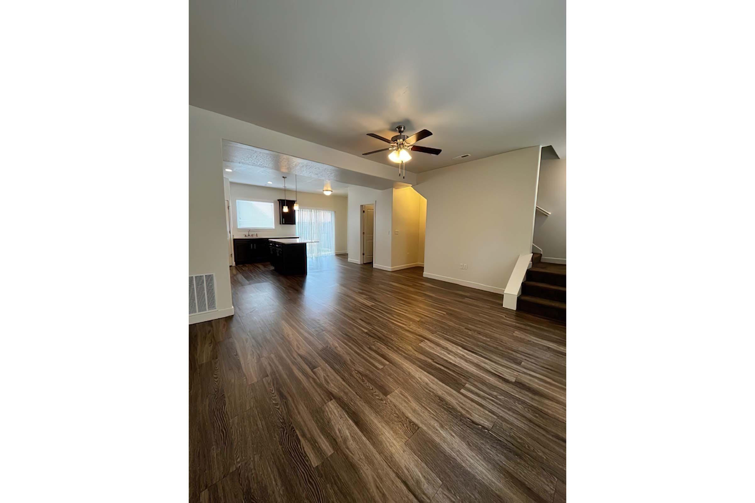 Spacious interior view of a modern living area featuring hardwood flooring, a ceiling fan, and an open layout. The kitchen is visible in the background with contemporary cabinetry, and there are stairs leading to an upper level. Natural light floods the space through large windows.