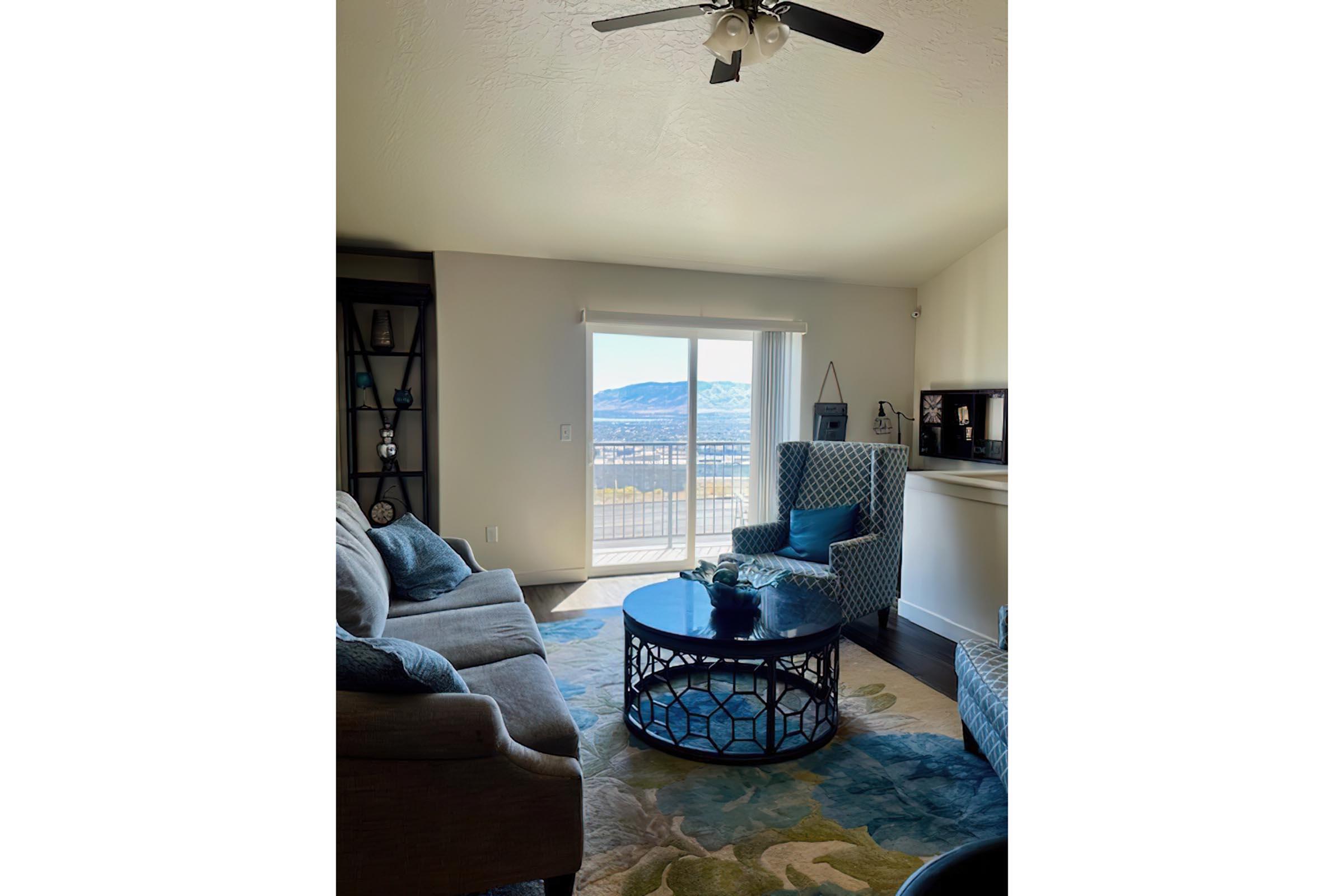 A bright living room featuring a comfortable gray sofa, two patterned blue chairs, and a round coffee table. Large windows allow natural light and offer a view of mountains outside. The room is decorated with colorful rugs and home accents, creating a cozy atmosphere.