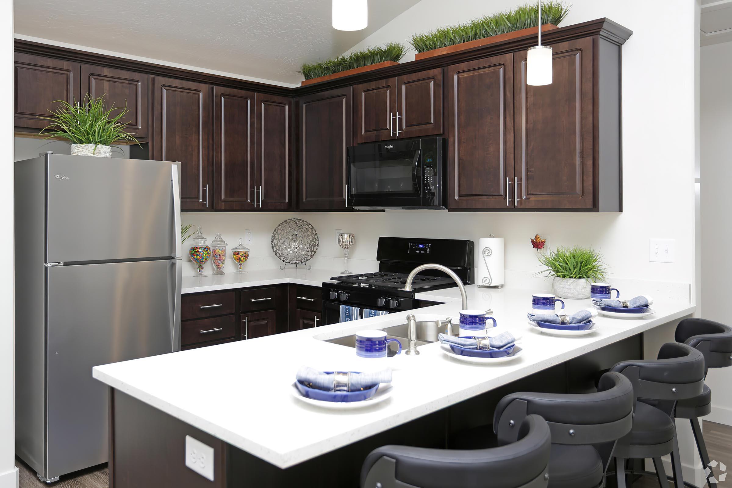 Modern kitchen featuring dark wooden cabinets, stainless steel refrigerator, black stove, and sleek white countertops. The dining area has a bar with black stools and a set table adorned with blue plates and utensils. Decorative plants add a touch of greenery to the space.