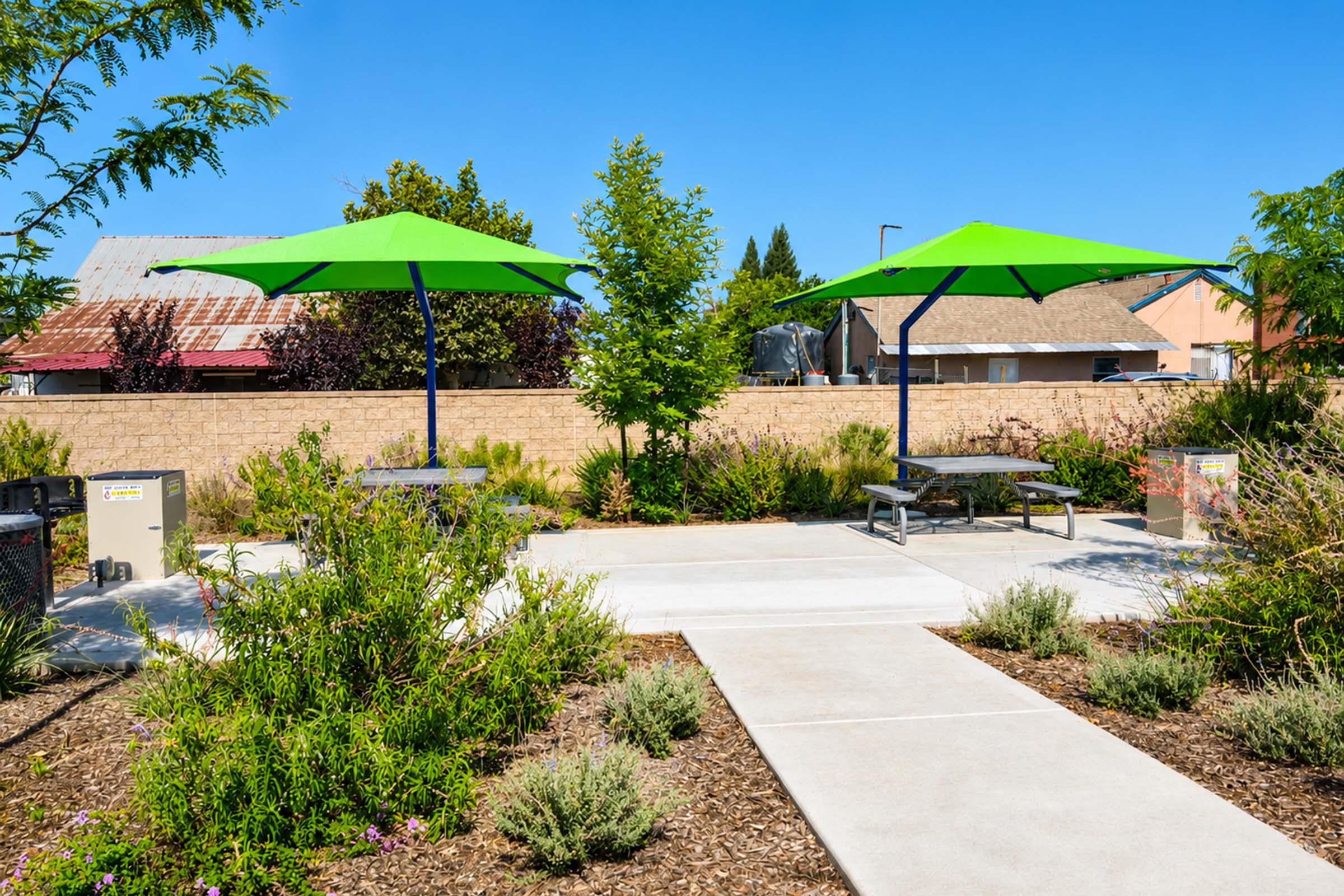 Two green umbrellas provide shade over picnic tables in a landscaped park area. The background features a wall, trees, and a building. A paved path leads through the greenery, with various plants and shrubs surrounding the seating area. The sky is clear and blue, indicating a sunny day.