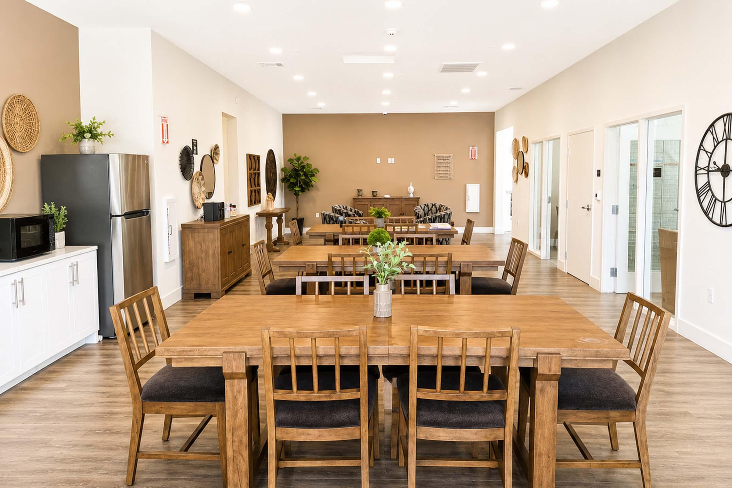 A modern common area featuring multiple wooden dining tables with chairs, a kitchenette with a refrigerator and microwave, and a cozy seating area with plants. The walls are light-colored, accented with decorative items, creating a welcoming and spacious atmosphere.