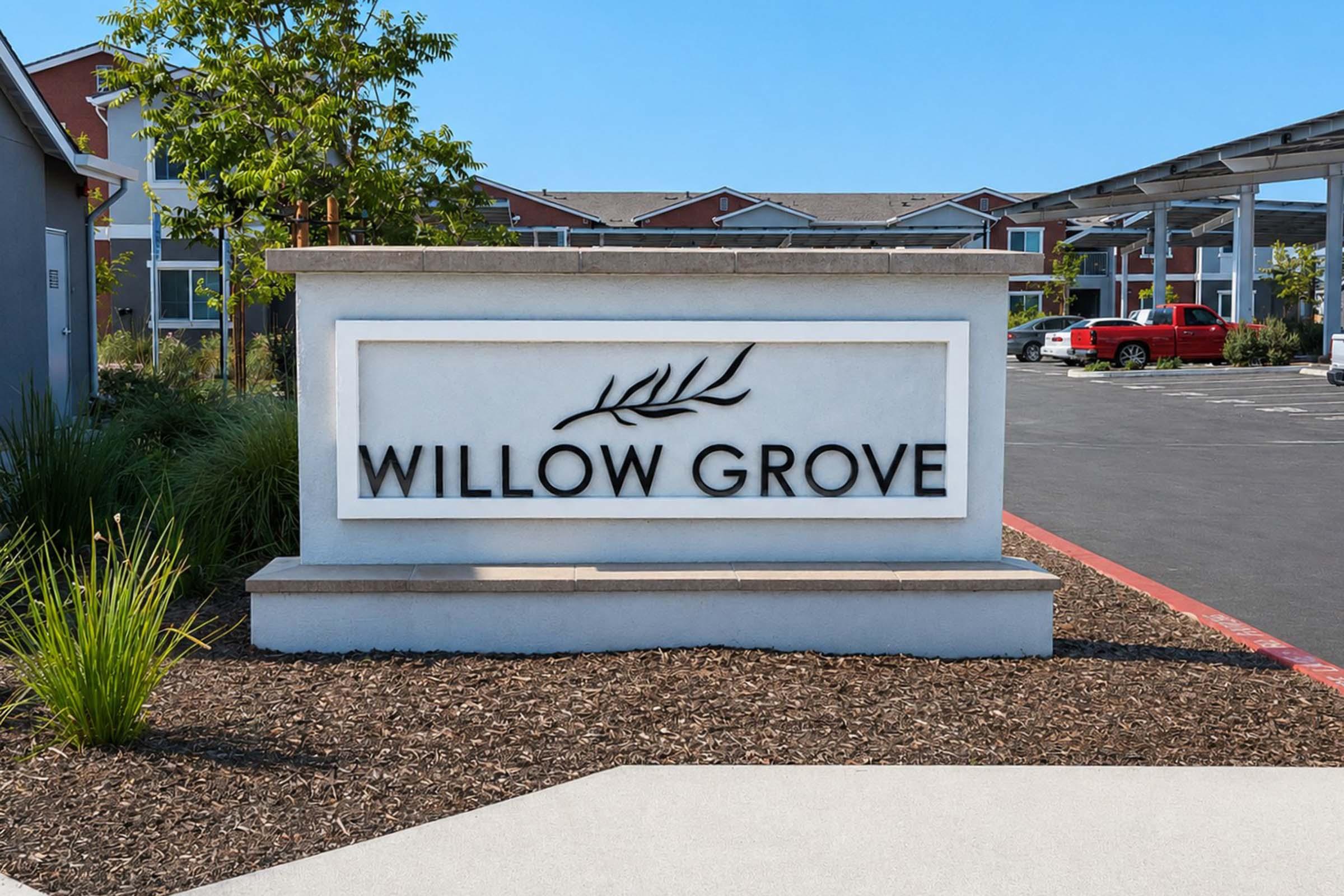 Sign for "Willow Grove" featuring a decorative leaf design, set against a landscaped backdrop. The sign is prominently displayed in a parking area, with modern buildings visible in the background and clear blue skies overhead.