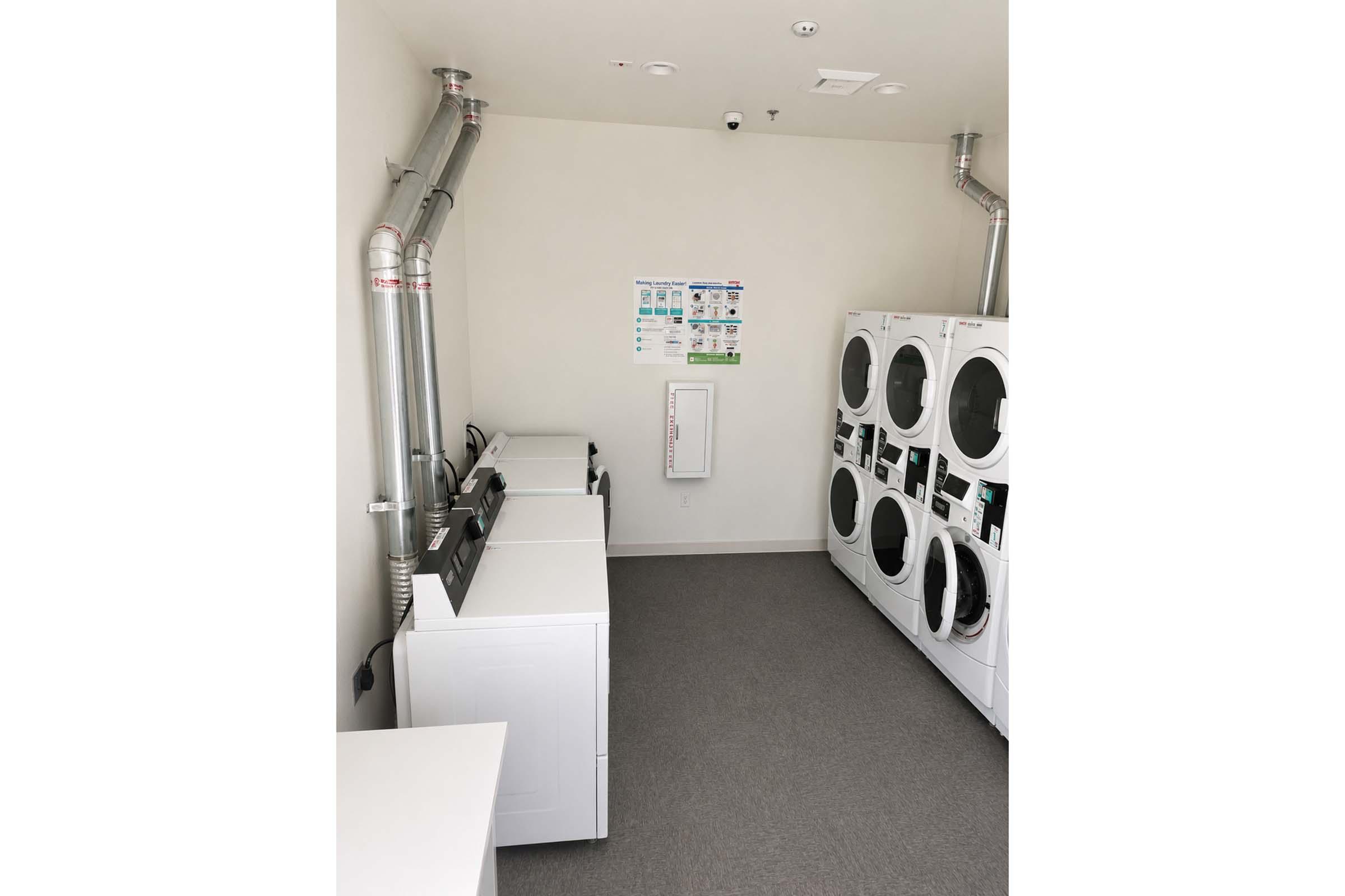 A clean, well-lit laundry room featuring multiple washing machines and dryers arranged against the wall. There are metal ventilation pipes above, and a bulletin board with laundry-related information on the wall. The floor is covered with gray carpeting, contributing to a modern and functional space.