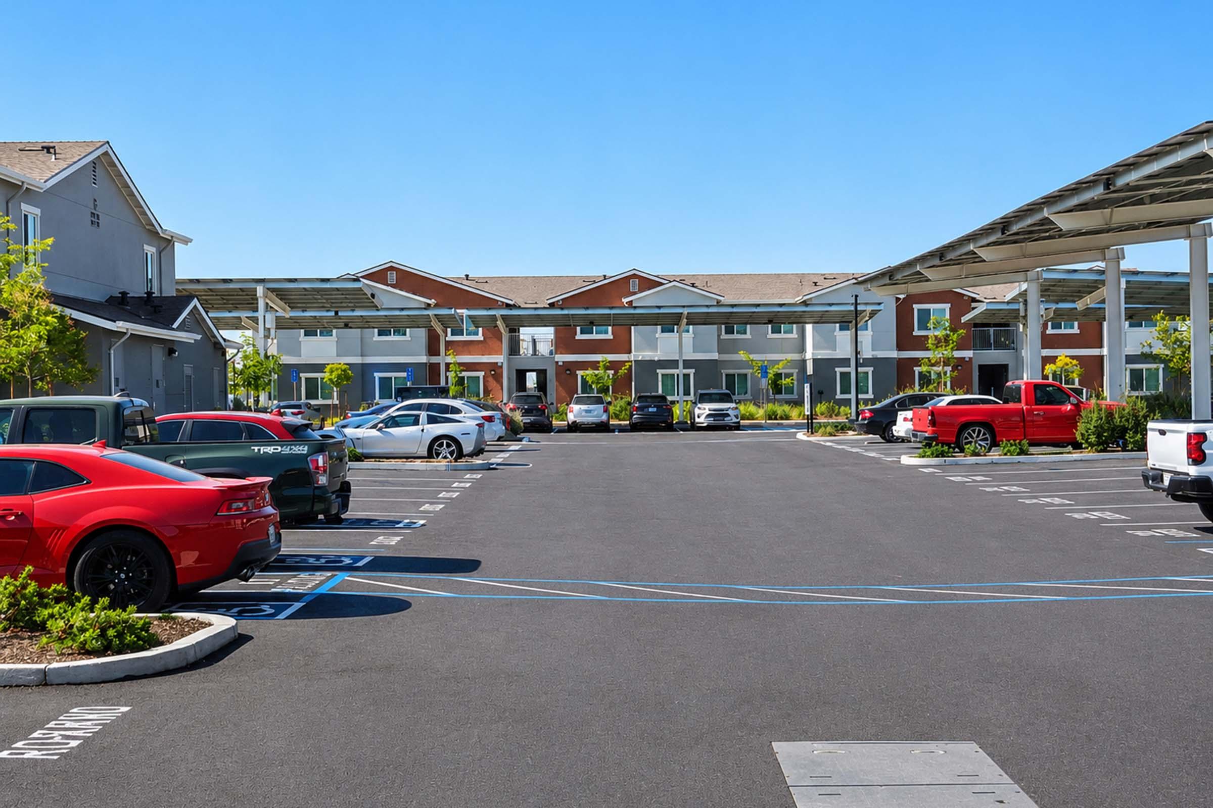 A view of a parking lot with various cars, including a red sports car and a red pickup truck. In the background, there are two-story apartment buildings with colorful facades. The sky is clear and blue, and there are small trees planted in the parking area, providing some greenery.
