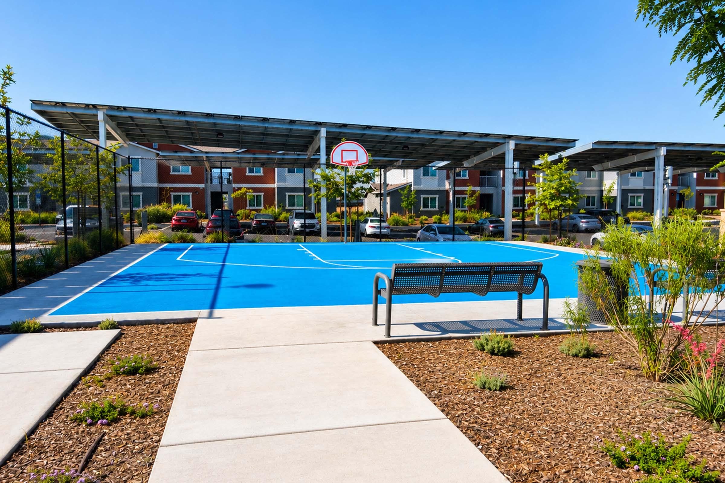 A vibrant blue basketball court with a hoop, surrounded by green plants and modern residential buildings. The court is partially shaded by solar panels, and there is a bench nearby for spectators. The scene is set under a clear blue sky, creating a lively outdoor sports environment.