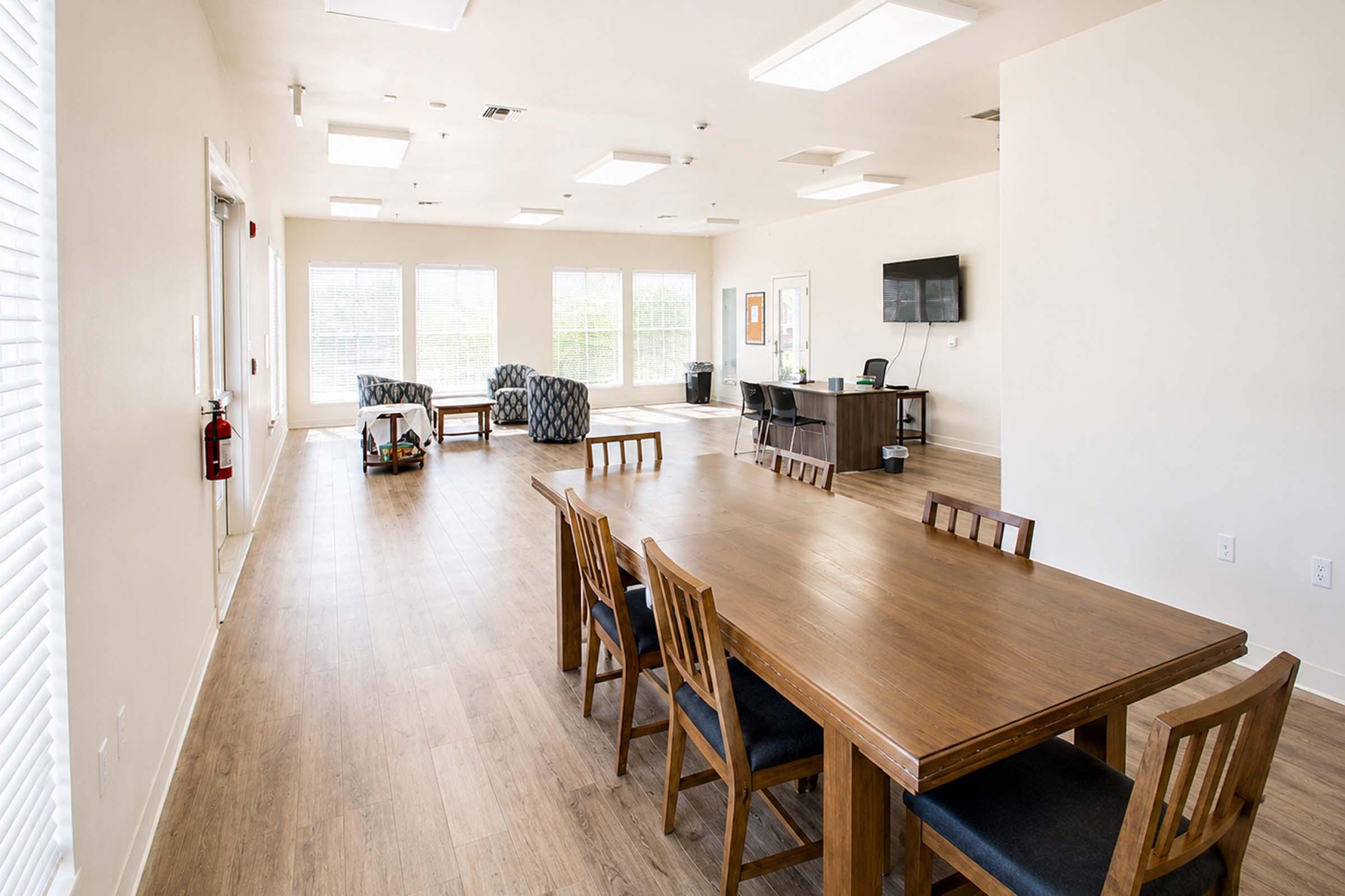 A spacious interior room with large windows allowing natural light, featuring a long wooden dining table with chairs, two patterned armchairs in the corner, a desk with a monitor, and a fire extinguisher mounted on the wall. The flooring is light wood, creating a warm and inviting atmosphere.