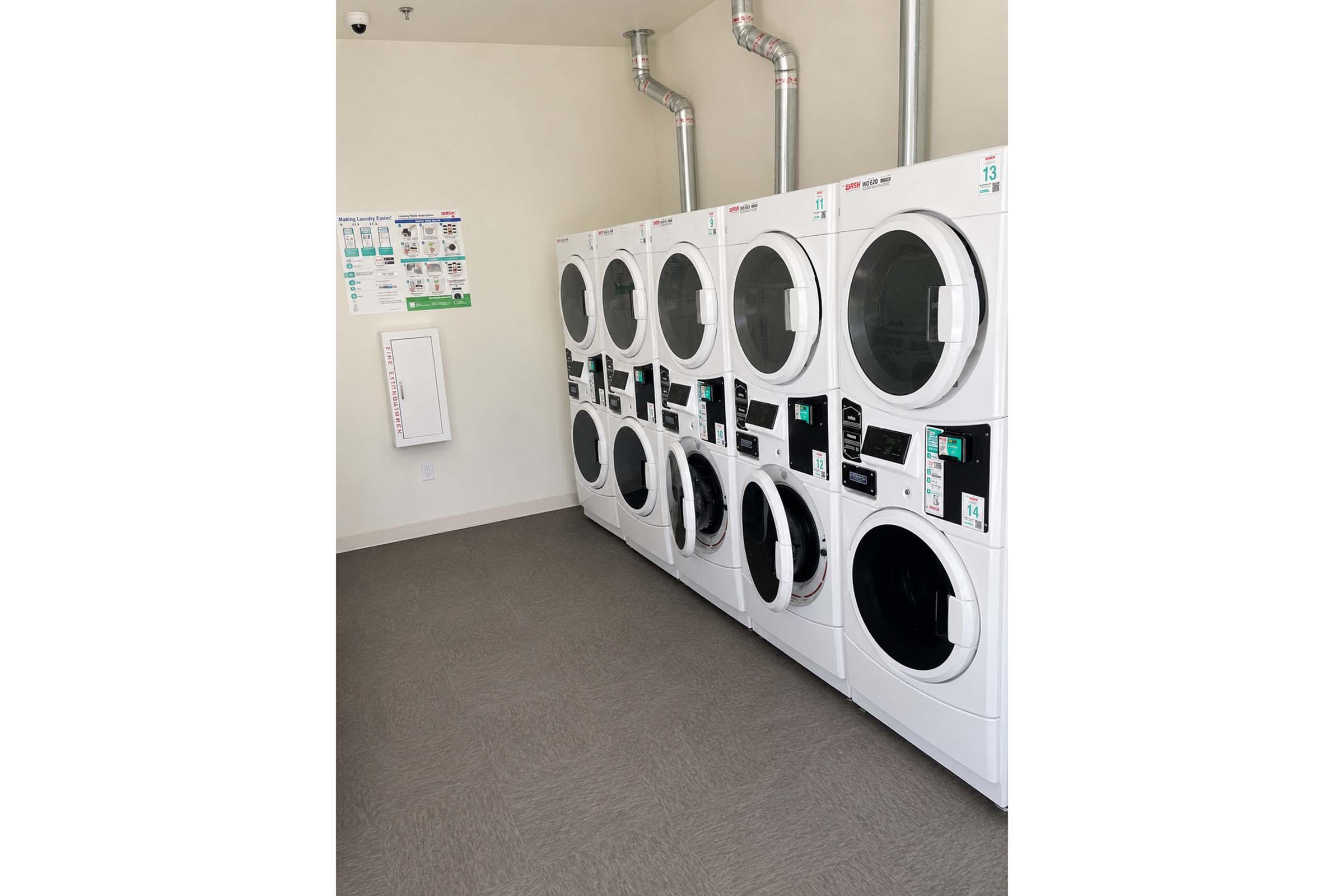A row of modern, front-loading washing machines and dryers in a well-lit laundry room. The appliances are white, with digital displays and coin slots. A wall-mounted information board is visible, and the floor is covered with gray carpeting.