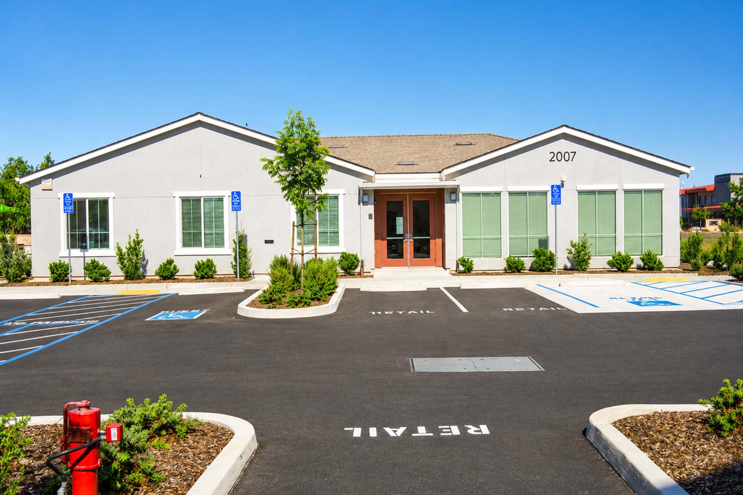 A modern single-story building with a light gray exterior and a pitched roof, featuring large windows and a centered entrance. The surrounding area has designated parking spaces labeled "RETAIL" and landscaped greenery. The building number "2007" is prominently displayed above the entrance.