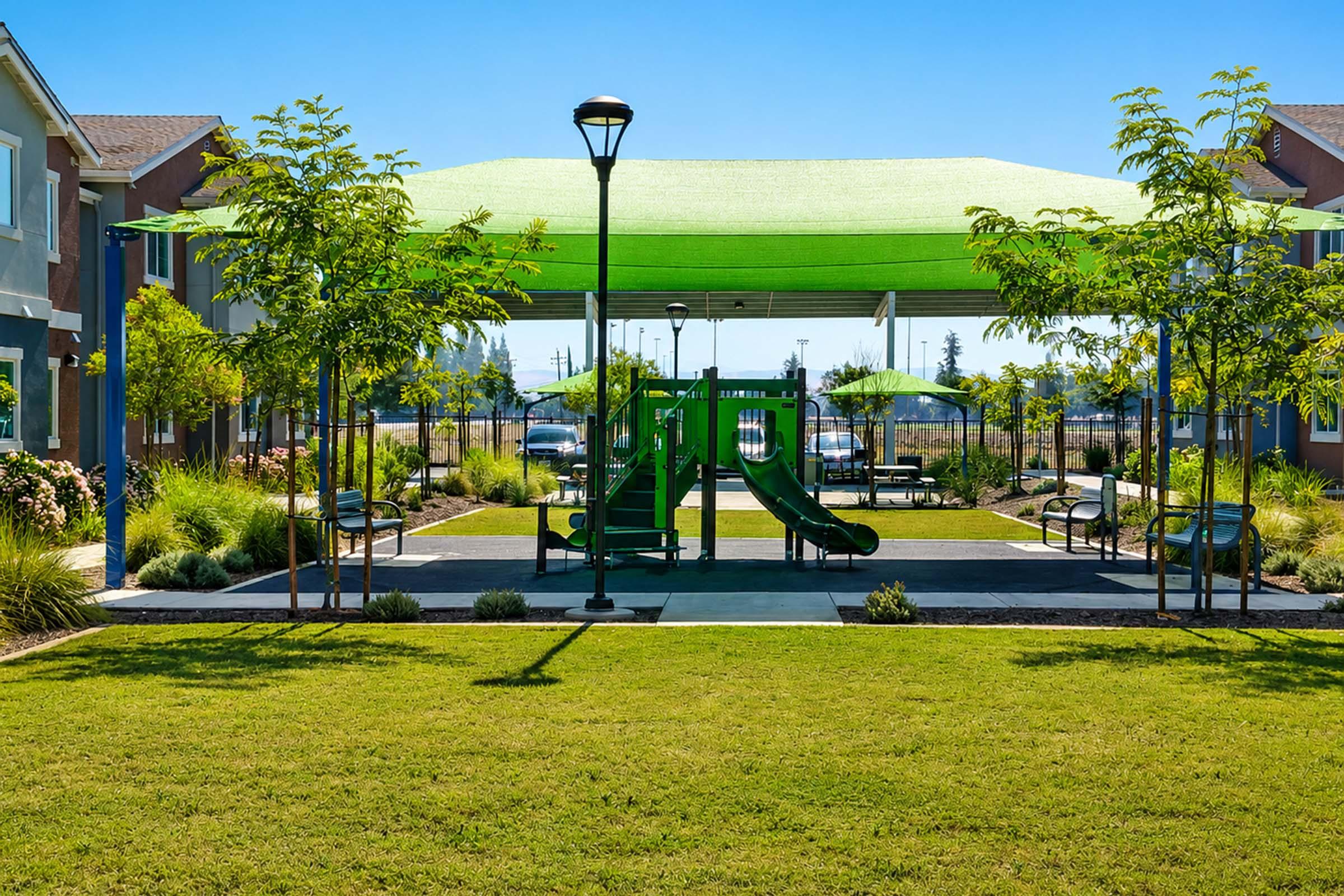 A shaded playground area featuring a green slide under a large canopy. Surrounding the playground are benches, grassy spaces, and well-maintained landscaping with small trees. Residential buildings are visible in the background, creating a bright, inviting outdoor space.
