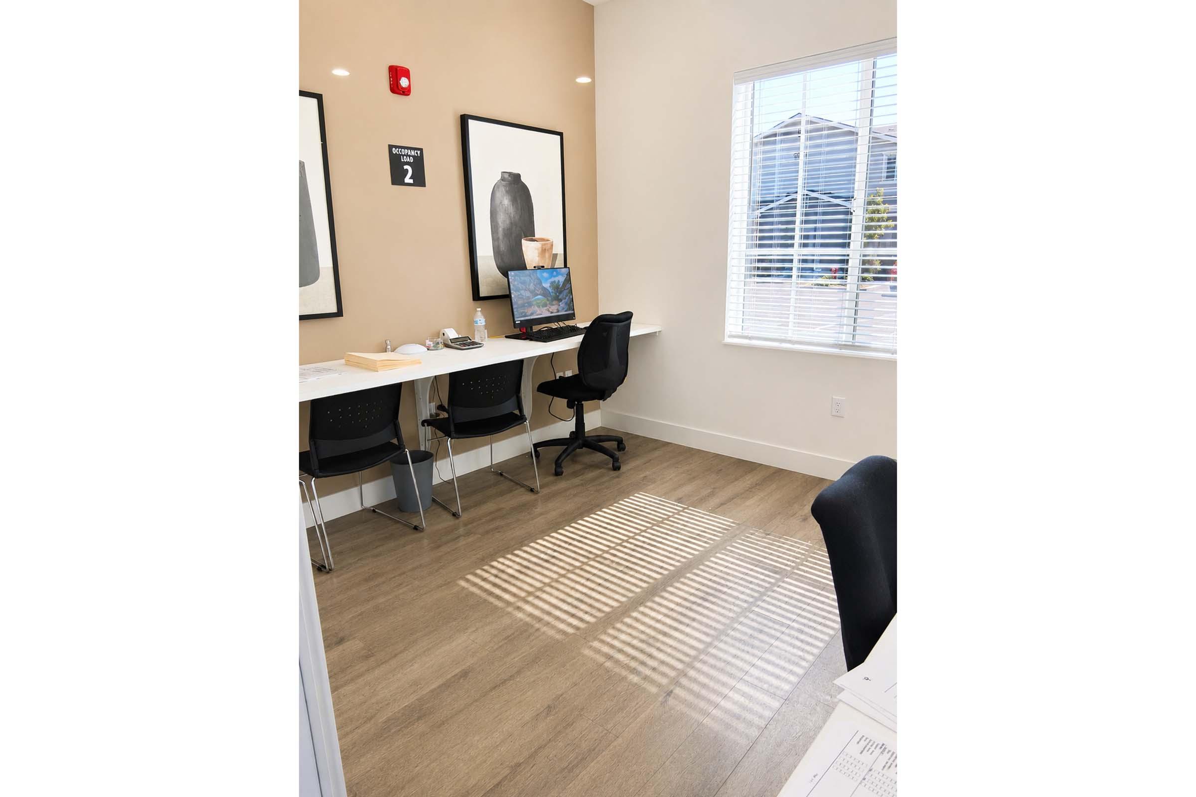 A small office space featuring a desk with a computer and a chair, located near a window. The walls are painted in a soft beige color, adorned with two large framed images of vases. Sunlight casts shadows on the wooden floor, creating a warm atmosphere. There’s a visible sign labeled "2" on the wall.