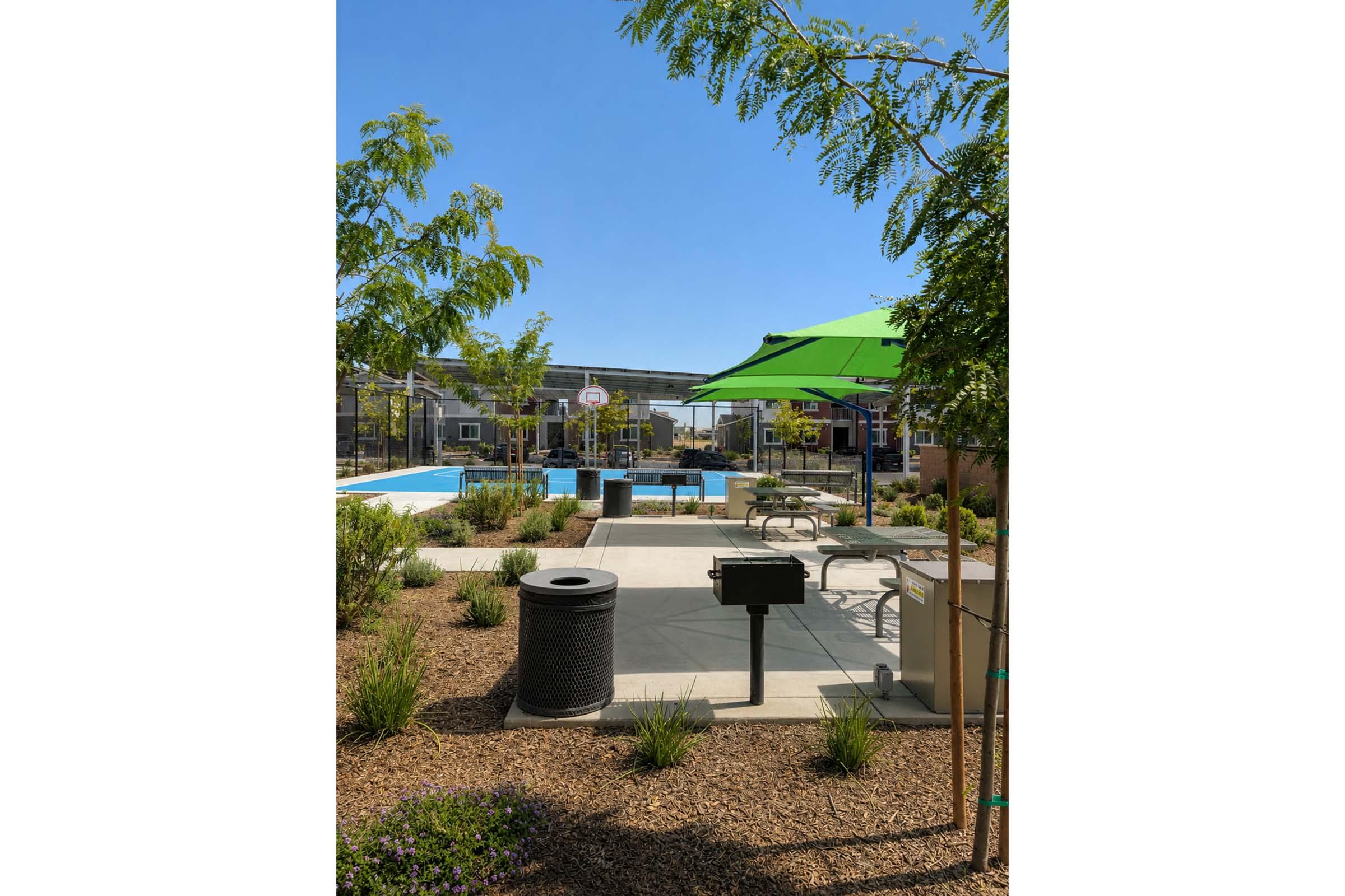 A sunny outdoor area featuring a swimming pool surrounded by lounge chairs and picnic tables. Green trees and plants decorate the space, and a charcoal grill is nearby, along with green umbrellas providing shade.