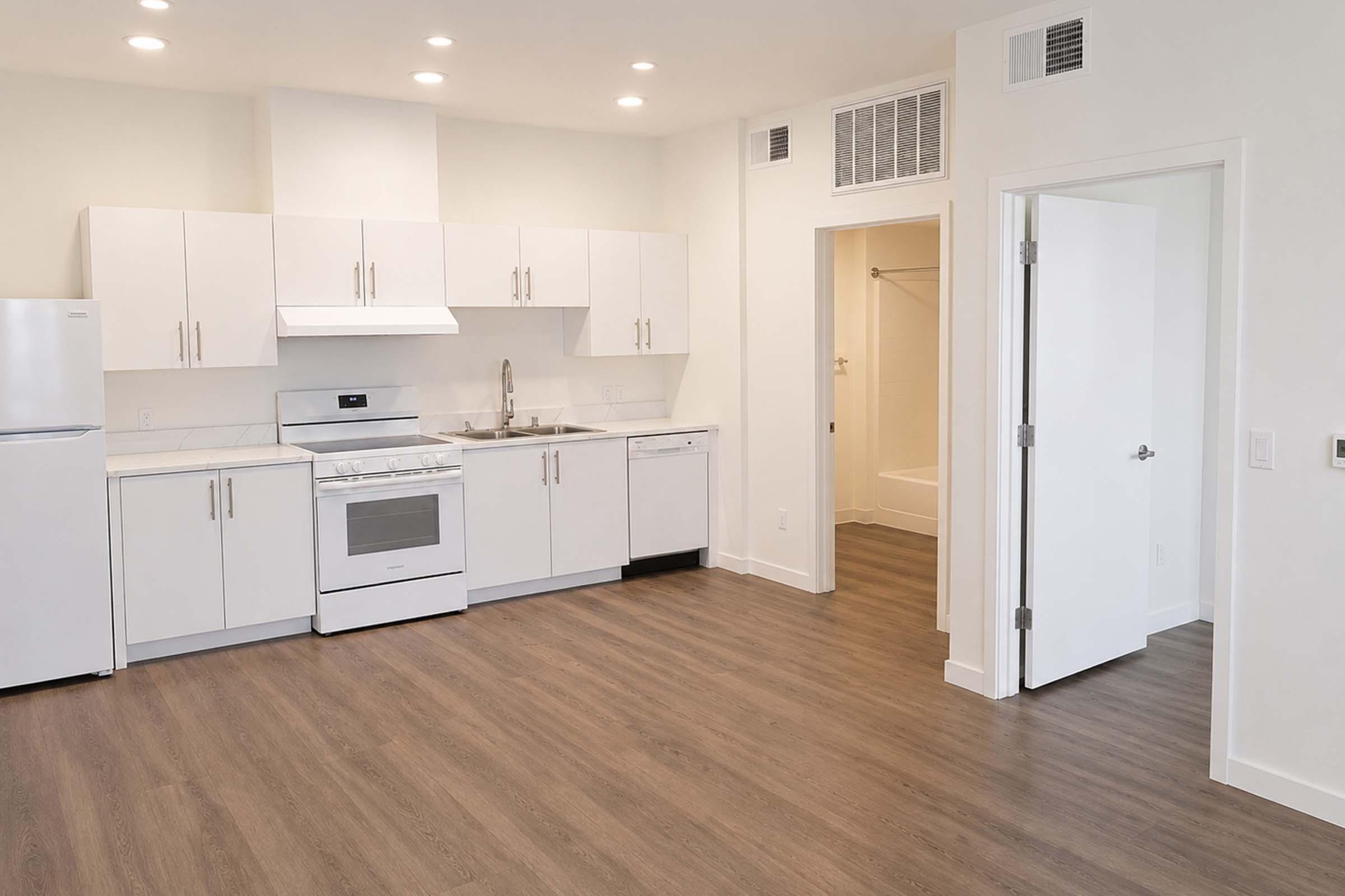 A modern kitchen featuring white cabinetry, a stainless steel oven, refrigerator, and dishwasher. The space has a light-colored backsplash and wood laminate flooring. There's an open layout with a doorway leading to a bathroom, enhancing the room's spacious feel with ample natural light from overhead fixtures.